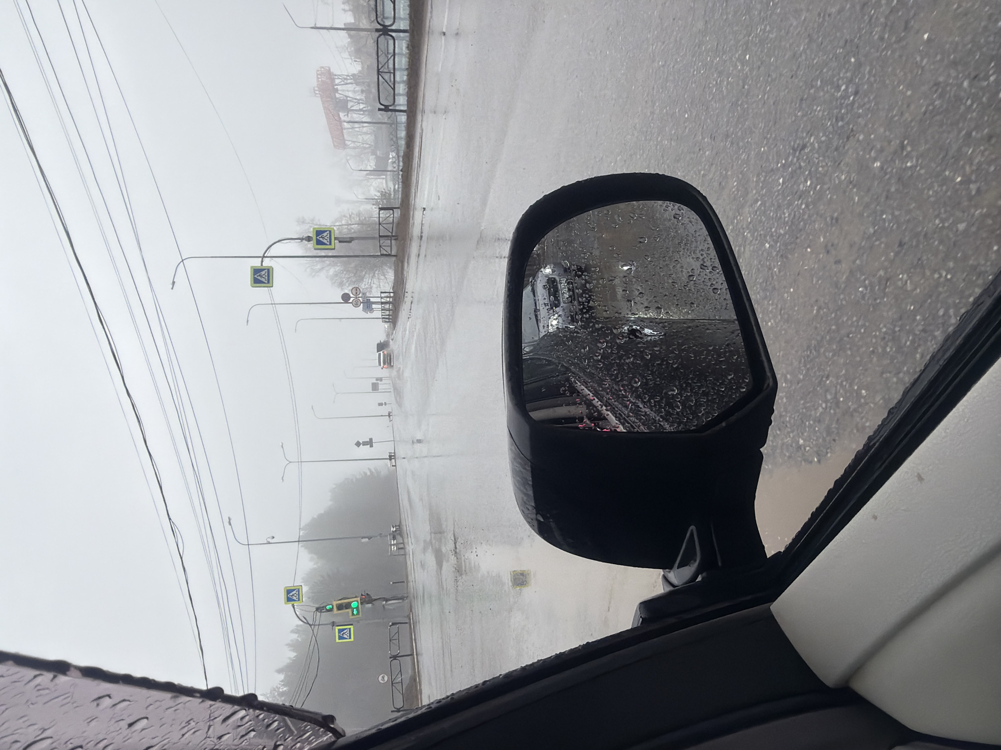 The image is taken from inside a car during heavy rain, with the car's side mirror prominently visible. Raindrops cover the mirror and the car's window. The side mirror reflects a view of the road behind, including another car with its headlights on. Outside the car, the road is wet and reflects the overcast sky. Streetlights and pedestrian crossing signs line the road. In the distance, a large metal structure, possibly a crane or industrial framework, is visible through the mist and rain. The image appears to be taken in Chaikovsky, Russia, as indicated by the location on the road and the architecture. The environment is bleak and wet, suggesting a typical rainy day in an urban area. There is no visible evidence of people.