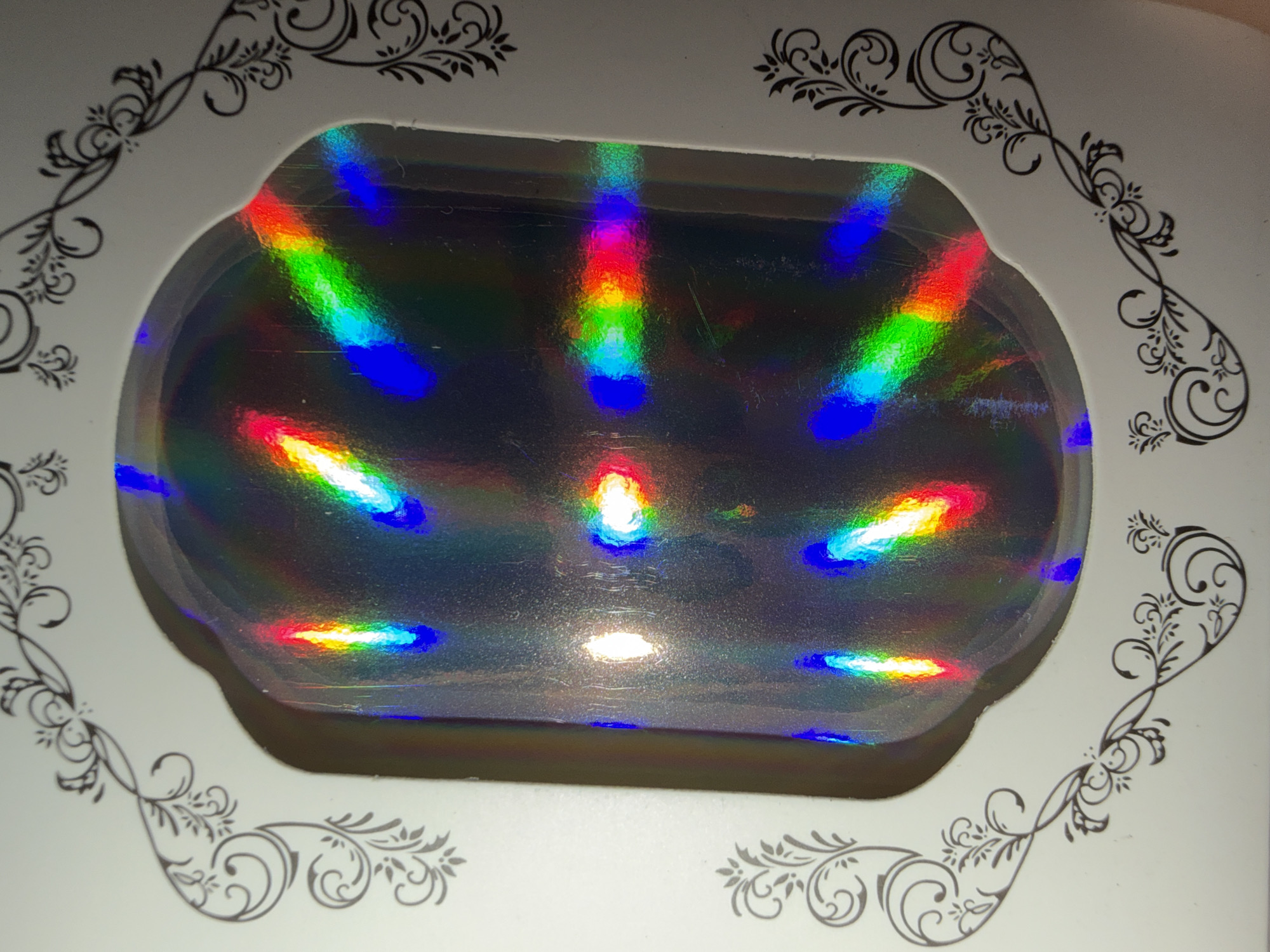 This is a close-up, top-down, eye-level photograph of a white box with decorative black scrollwork on its edges. The box has a clear, oval-shaped window through which a holographic or iridescent surface is visible. This surface reflects light in a rainbow spectrum, creating distinct bands of color across its entirety. These colors include vibrant reds, oranges, yellows, greens, blues, and purples. The light reflections are not uniform, appearing as streaks and patches of color, some brighter than others, suggesting a light source from above. The holographic surface appears slightly textured with minor scratches or imperfections visible. The overall impression is one of light, color, and a decorative, possibly gift-like, item. The setting appears to be indoors, likely a home or a retail environment, given the lighting and the nature of the object. No people are visible in the image.