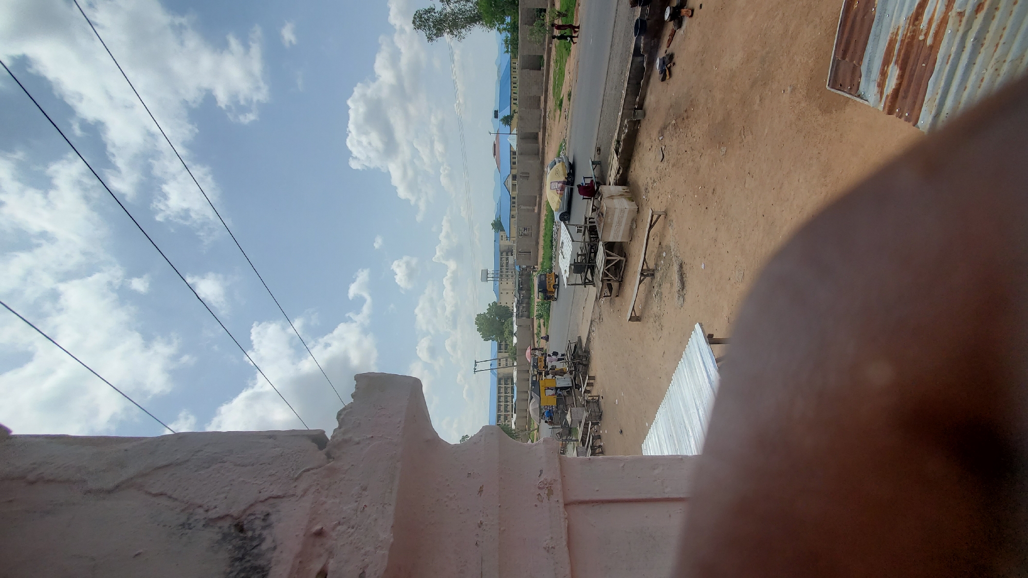 In this outdoor, daylight scene, a dirt road runs horizontally through the frame. Several open-air market stalls with corrugated metal roofs are visible along the road. A light-colored car with a yellow cover is parked on the side of the road, and a yellow motorized tricycle is driving on the road. In the background, there are buildings with blue roofs. The sky is blue with scattered white clouds, and several power lines are strung across the upper portion of the image. The foreground is partially obscured by a blurred, brown, organic shape, possibly a person's hand or arm, and a section of a corrugated metal roof. The scene appears to be in a semi-developed area with a mix of commercial and residential structures.