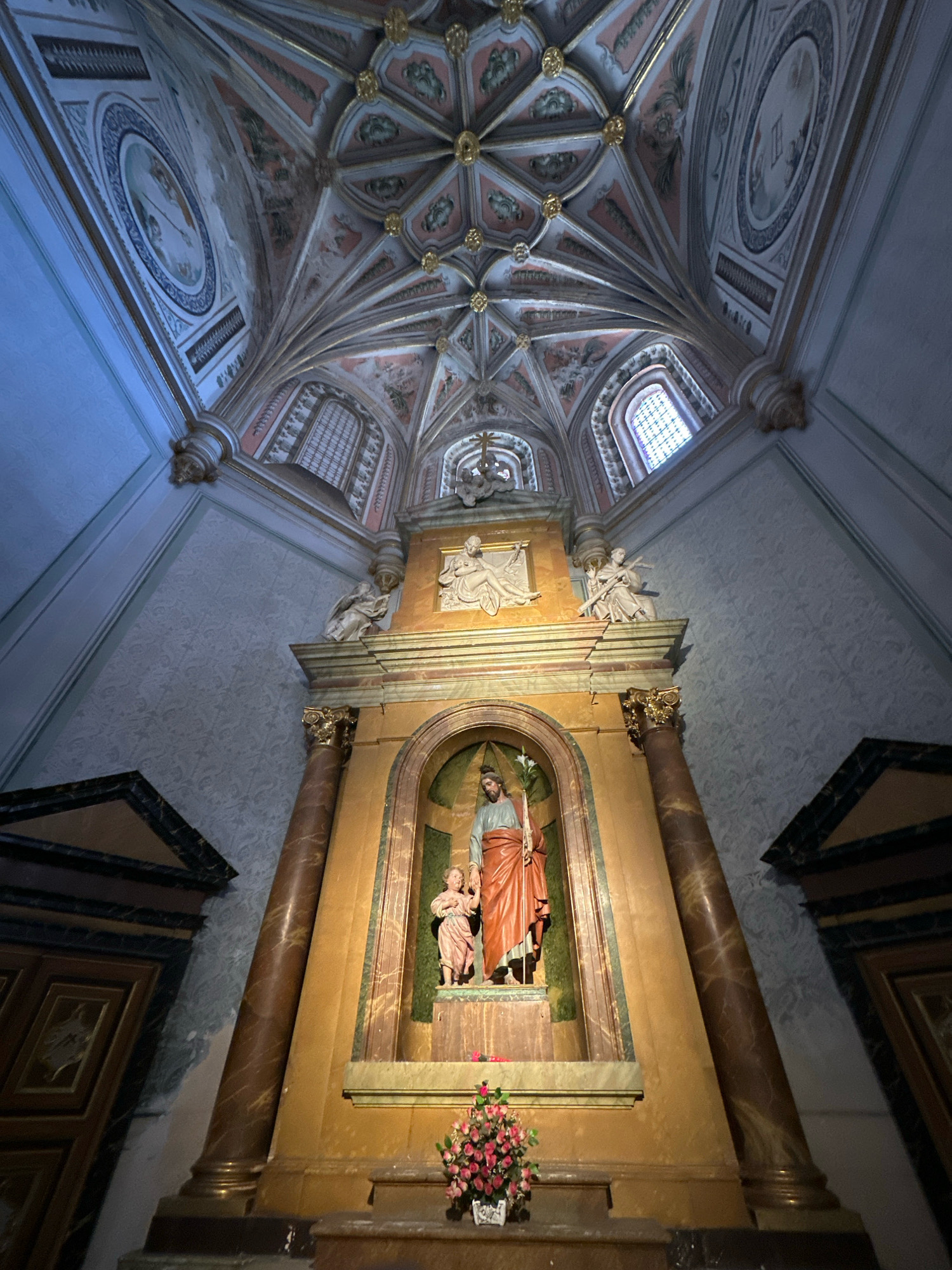 The image shows the interior of a building with a high, vaulted ceiling decorated with intricate geometric patterns and painted murals. The ceiling features gold accents and ribbed vaults. Below the ceiling, a large altar is visible, dominated by a statue of Saint Joseph holding the child Jesus. The statue is enclosed within an arched niche, and the altar area is adorned with marble columns and decorative architectural elements. To the left and right of the altar are large, dark wooden doors with decorative carvings. Floral arrangements are placed at the base of the altar. The walls are covered in a textured wallpaper with a subtle pattern. The overall impression is of a richly decorated religious building, likely a church or chapel. The location is specified as the city of La Lastrilla, Spain.