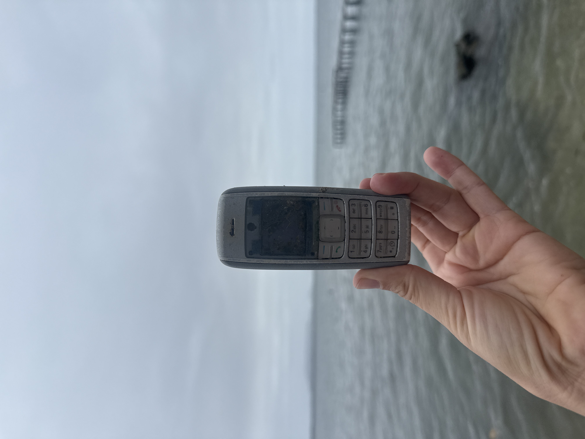 A weathered, light gray Nokia feature phone is held in the foreground by a person's hand. The phone's screen is dark and smudged, and the casing appears dirty. Behind the phone, a calm, gray body of water stretches to the horizon under an overcast sky. In the middle distance, a dilapidated wooden pier extends into the water. The scene suggests a location near a coast or large body of water, possibly in a temperate climate given the cloudy sky.