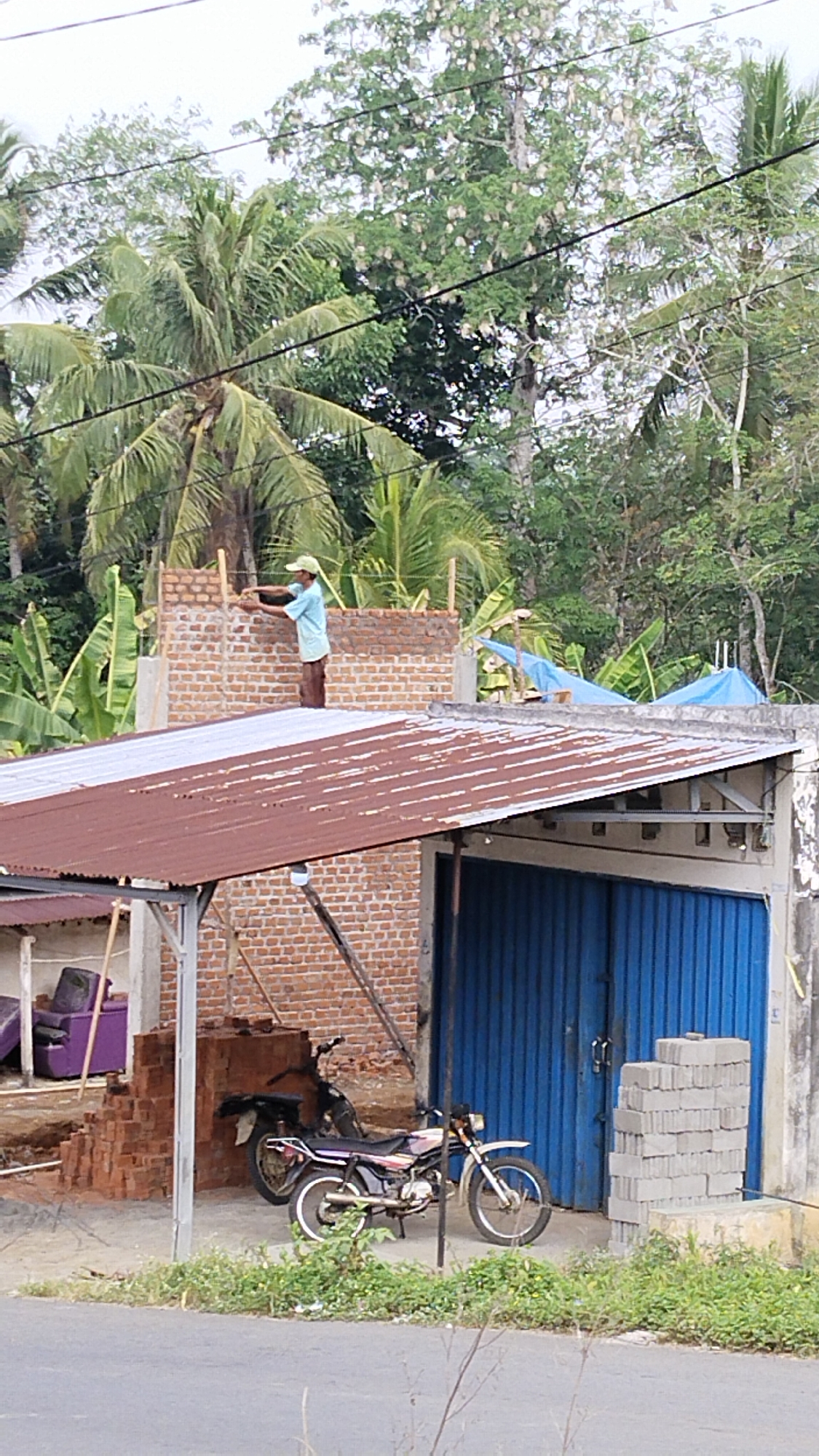 The image depicts a construction site in Margatunggal, Indonesia. A construction worker, wearing a light blue t-shirt and brown trousers, is positioned on top of a partially constructed brick wall, appearing to be laying bricks. The wall is under construction, supported by wooden scaffolding. In the foreground, a portion of a corrugated metal roof extends from a building. A blue metal garage door is visible on the right side of the building, and a stack of gray concrete blocks rests beside it. Two motorcycles are parked near the building, one in front of the other. A purple armchair is partially visible on the left side of the image, behind a pile of bricks. The background consists of lush green trees and palm trees, indicative of a tropical environment. Overhead power lines are also visible. The scene is outdoors, with natural lighting.