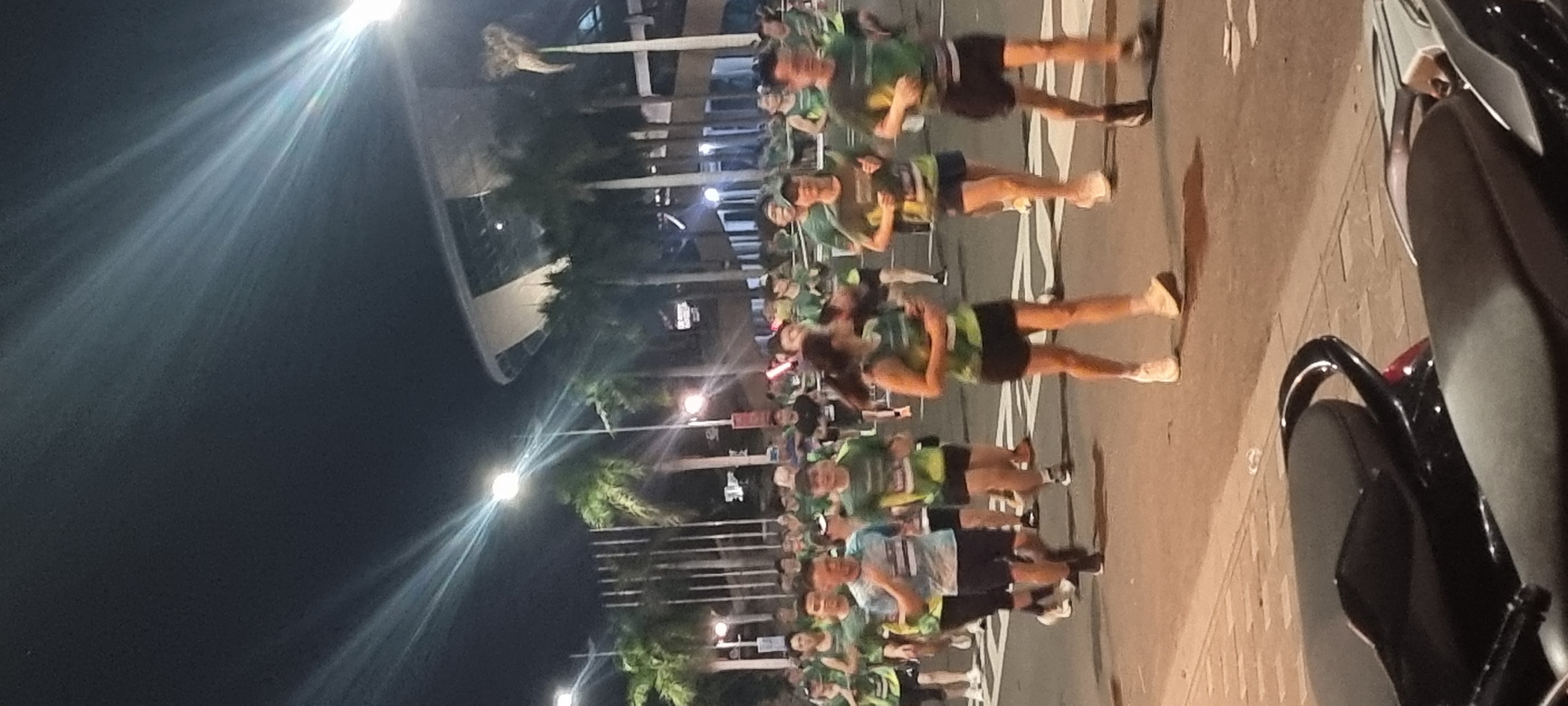 This is a full-shot, eye-level, vertical photograph of a night marathon in Đà Nẵng, Vietnam. The image captures a group of runners, mostly wearing green t-shirts and black shorts, in motion on a paved road. The runners are primarily of East Asian descent, with a few individuals who appear to be of European or mixed ethnicity. Some runners are focused on the race, while others are looking around.
In the background, tall palm trees line the street, and a modern, multi-tiered building with a curved roof is visible, suggesting an urban setting. Streetlights with bright, radiating beams cast light over the scene, creating streaks and lens flares that are prominent in the upper portion of the image. The road is marked with white lines, and the sidewalk on the left is paved with square tiles. In the foreground, the seats of two parked motorcycles are visible, indicating they are part of the street furniture or have been placed there for the event. The atmosphere appears to be energetic, with the light beams and the movement of the runners suggesting a lively event.