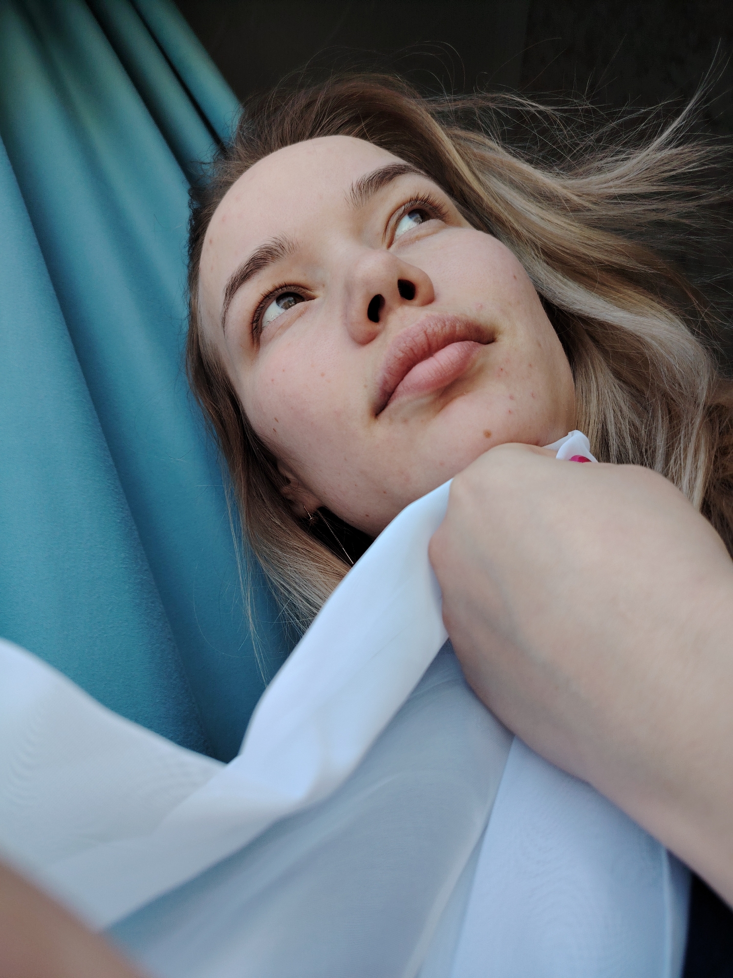 A young adult female with light brown and blonde-highlighted hair, fair complexion, and light-colored eyes is captured in a close-up, angled shot. She is looking upwards and slightly to the left, with her lips slightly parted. Visible on her face are several minor skin blemishes. Her right hand, featuring a fingernail with pink polish, is holding or gripping a piece of white, possibly sheer, fabric near her neck and upper chest area. The subject is wearing a dark garment, partially visible on the right. The immediate background to her left consists of a teal-colored curtain or drapery with vertical folds, suggesting an indoor setting within the city of Марковский, Russia. To her upper right, behind her hair, is a dark, indistinct background.