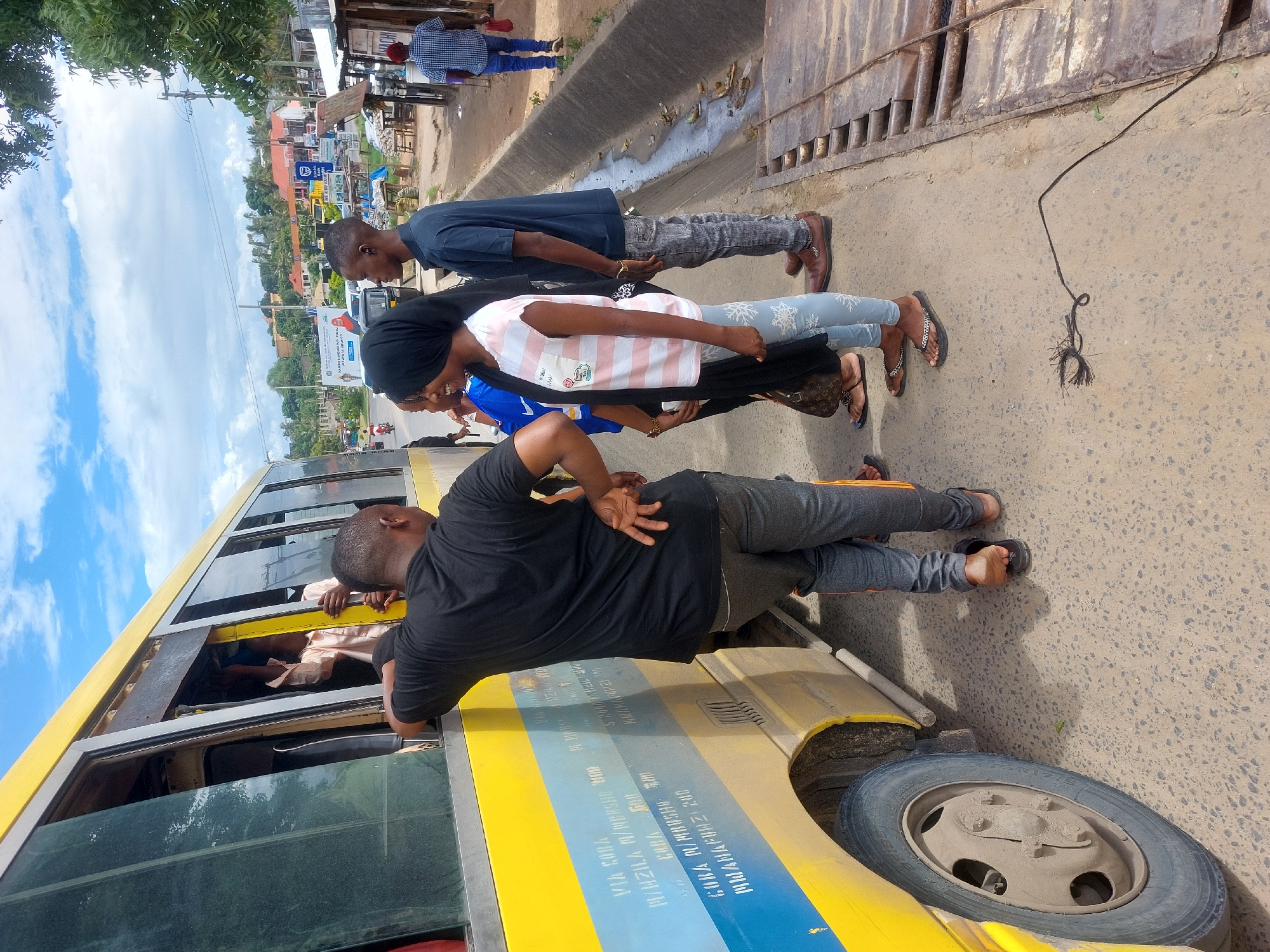 The image depicts a scene likely in Dar es-Salaam, Tanzania, indicated by the yellow commuter bus bearing destination names. Several people are standing next to the bus, appearing to interact with those inside. The bus displays destination names such as "VIA COBA" and fares in Tanzanian Shillings (TZS), such as "MWANAFUNZI 200," indicating student fares. The setting is an urban roadside with shops and other businesses visible in the background. A drainage ditch runs parallel to the road. The people are dressed in casual clothing.