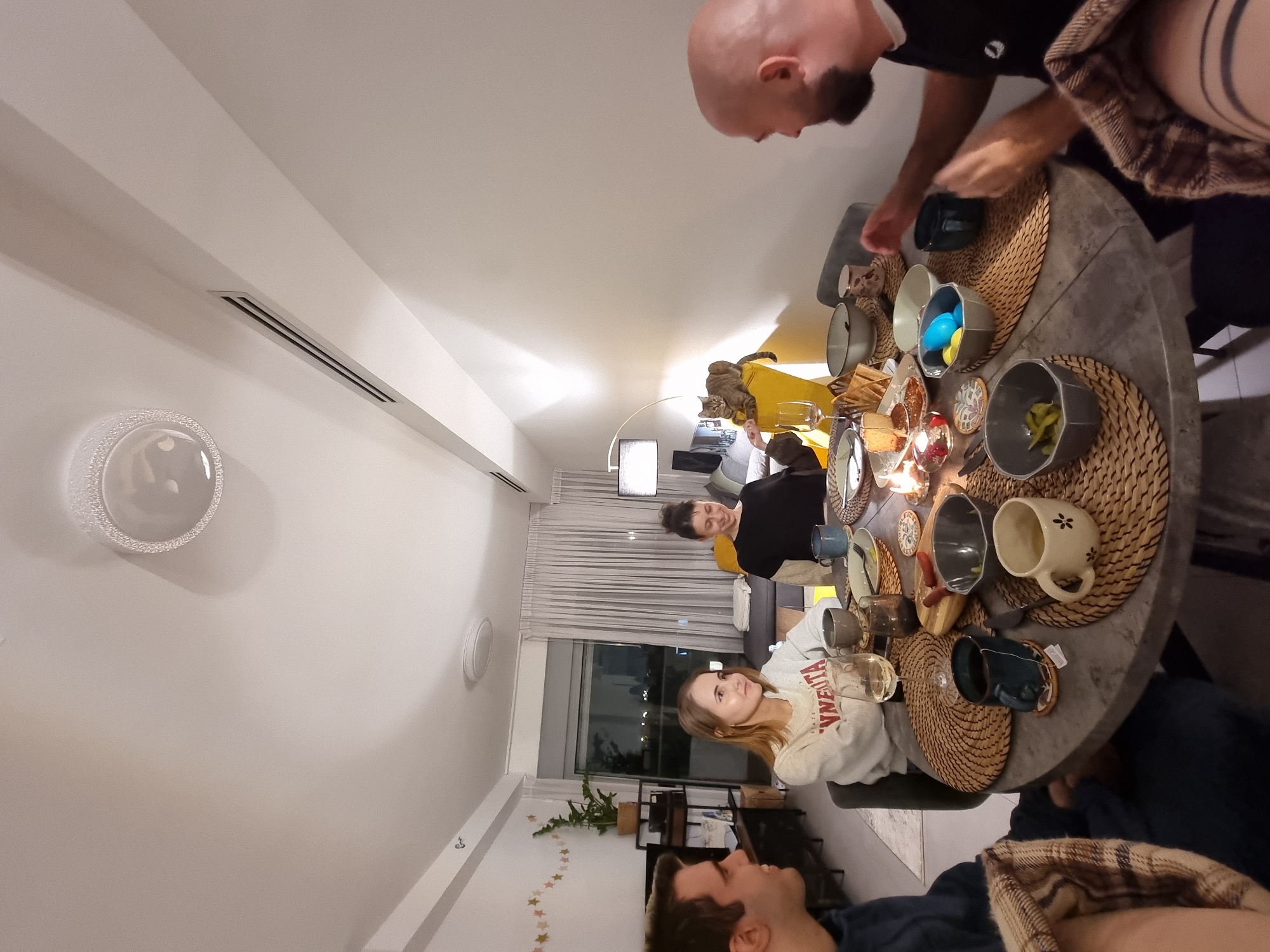 A wide-angle shot captures a gathering of people at a round table in what appears to be an apartment setting, likely in Ramat Gan, Israel. The focus is on the people around the table and the various objects on the table, including placemats, mugs, plates, bowls with food, glasses, and candles. People are interacting and some are eating or serving. The apartment interior includes a large window, curtains, plants, a decorative light fixture on the wall, and other furniture. A cat is seen resting on a yellow chair in the background. The perspective is tilted, which gives a slightly distorted view of the room's dimensions and the arrangement of objects.