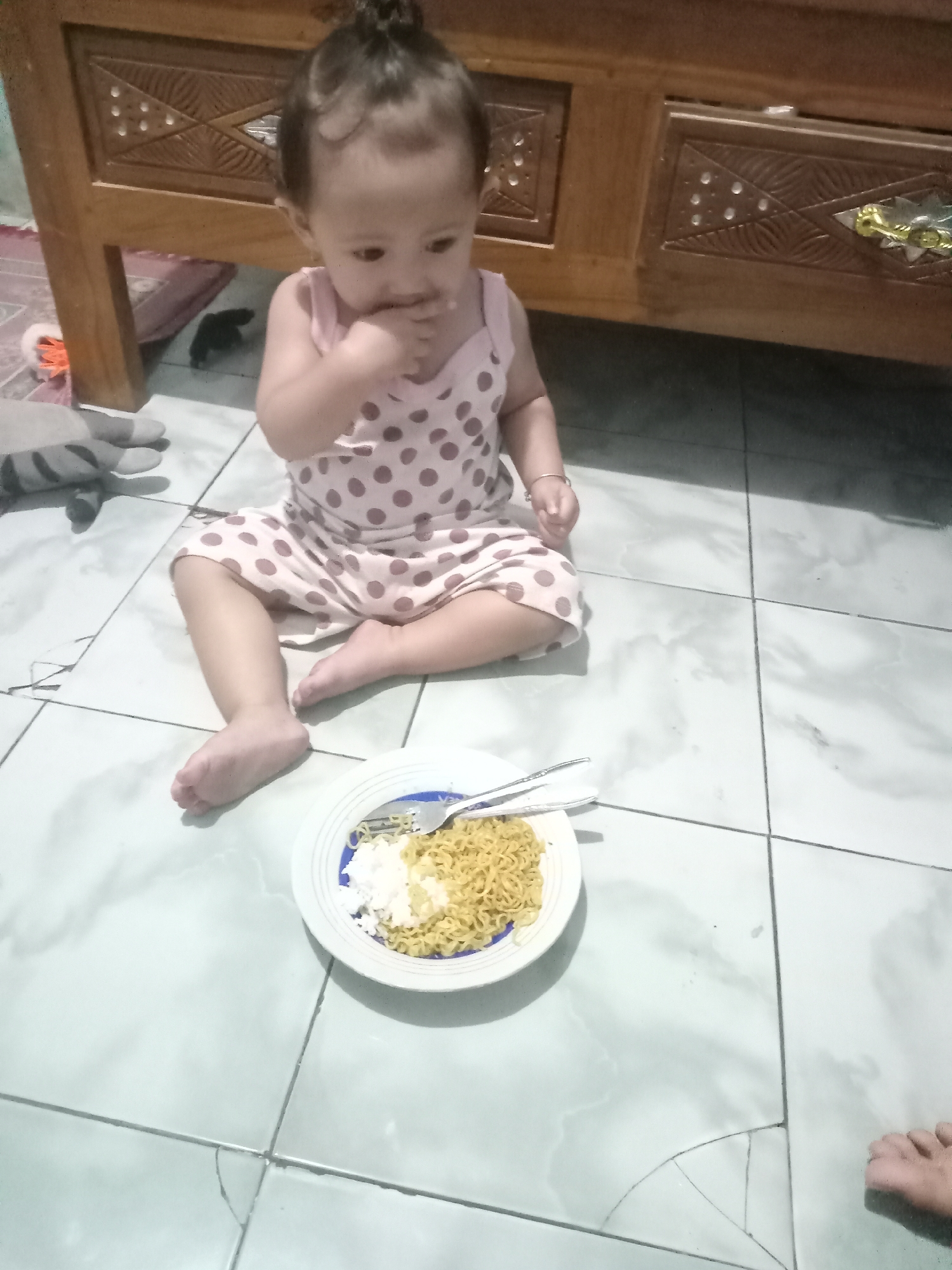 A young child wearing a light pink dress with brown polka dots is sitting cross-legged on a light grey tiled floor. The child has dark hair styled in a topknot and is holding their finger to their mouth. A white plate with a blue rim is positioned in front of the child, containing a serving of cooked noodles and a small portion of white rice. A fork and spoon rest on the plate.
Behind the child is a wooden dresser with decorative carved panels. Part of a grey plush toy is visible to the left of the child. The location is potentially Bungur, Indonesia.