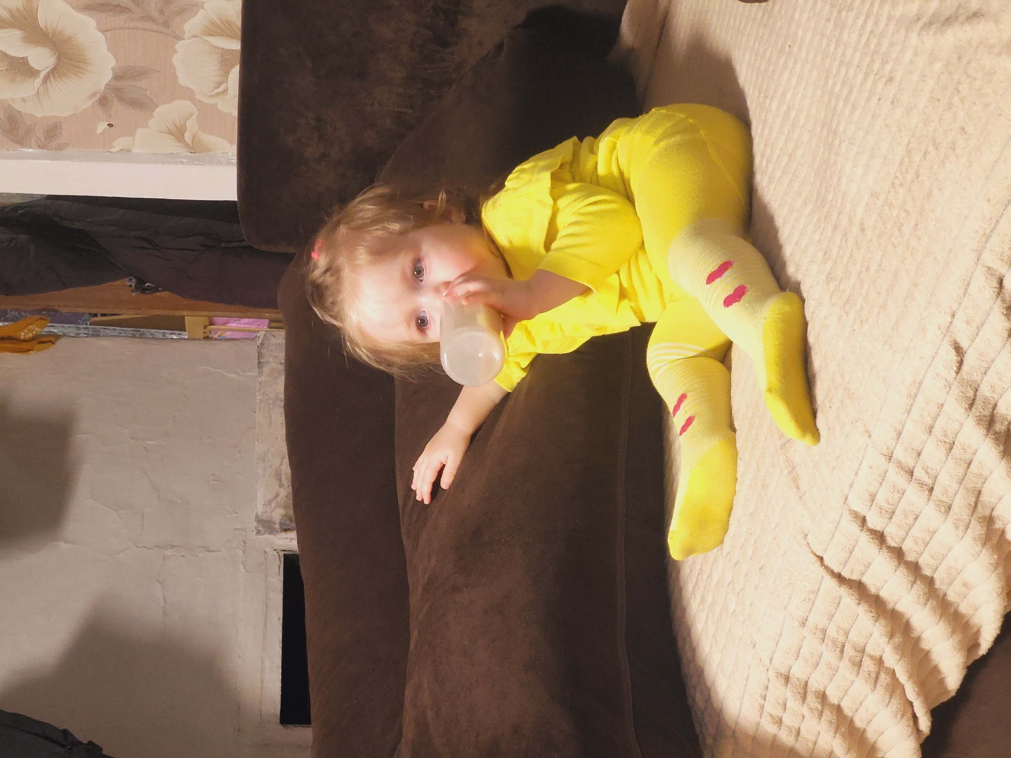 A young child wearing a yellow onesie and yellow socks with pink stripes on the lower leg is lying on a brown couch, drinking from a clear baby bottle. The couch is next to a beige blanket and in front of a wall with floral wallpaper. The location is Зипуново, Russia.