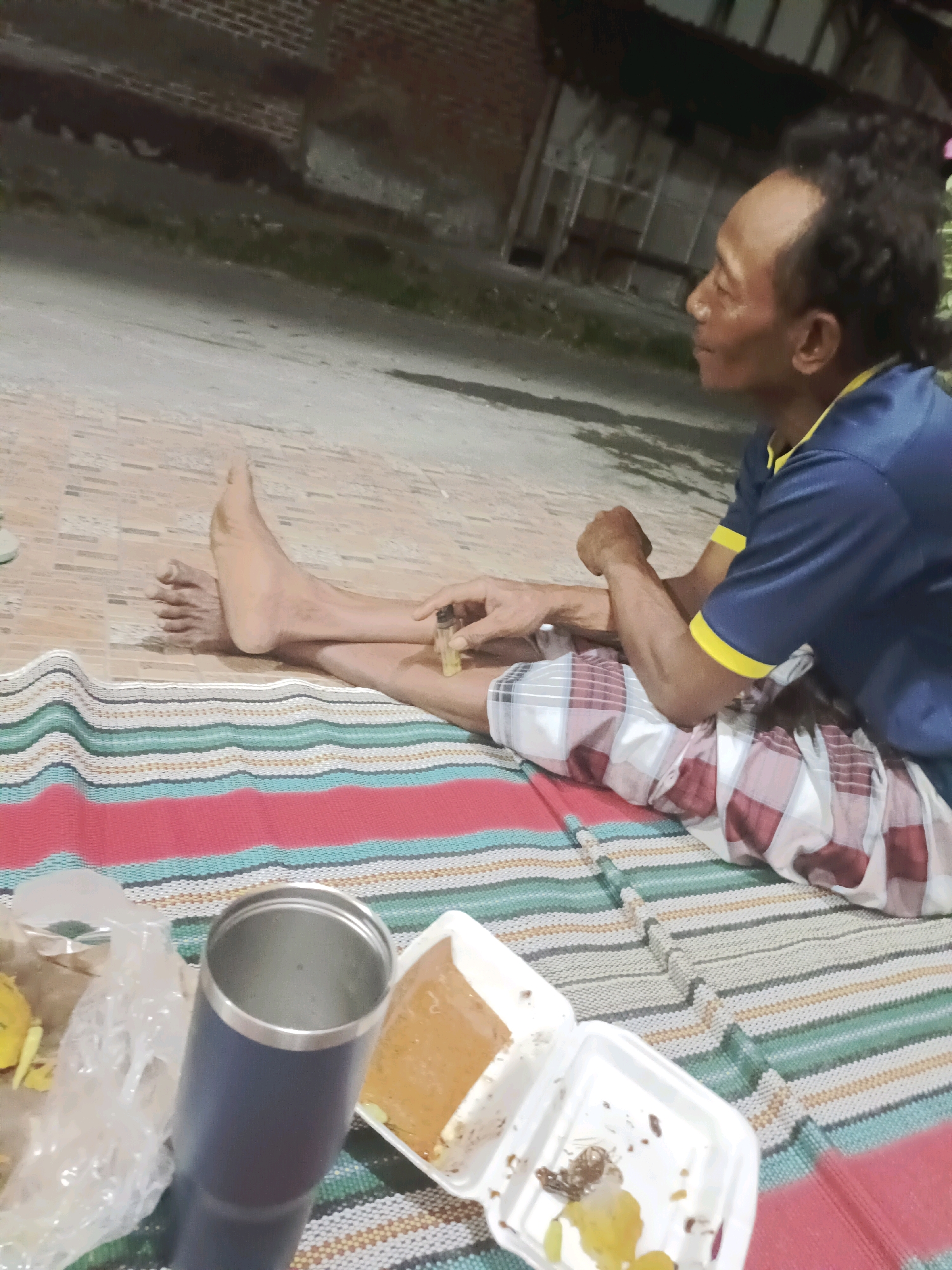 The image shows a man sitting on a striped mat outdoors, possibly in the city of Rancang Kencono, Indonesia. He is wearing a blue t-shirt with yellow trim and plaid shorts. His legs are crossed, and he is holding a small glass bottle in his right hand. In front of him, on the mat, are a dark blue insulated tumbler, a plastic bag with some yellow items inside, and a white takeout container with some food remnants. The background appears to be an outdoor area with a brick wall and a paved surface. The lighting suggests it is either dusk or nighttime.