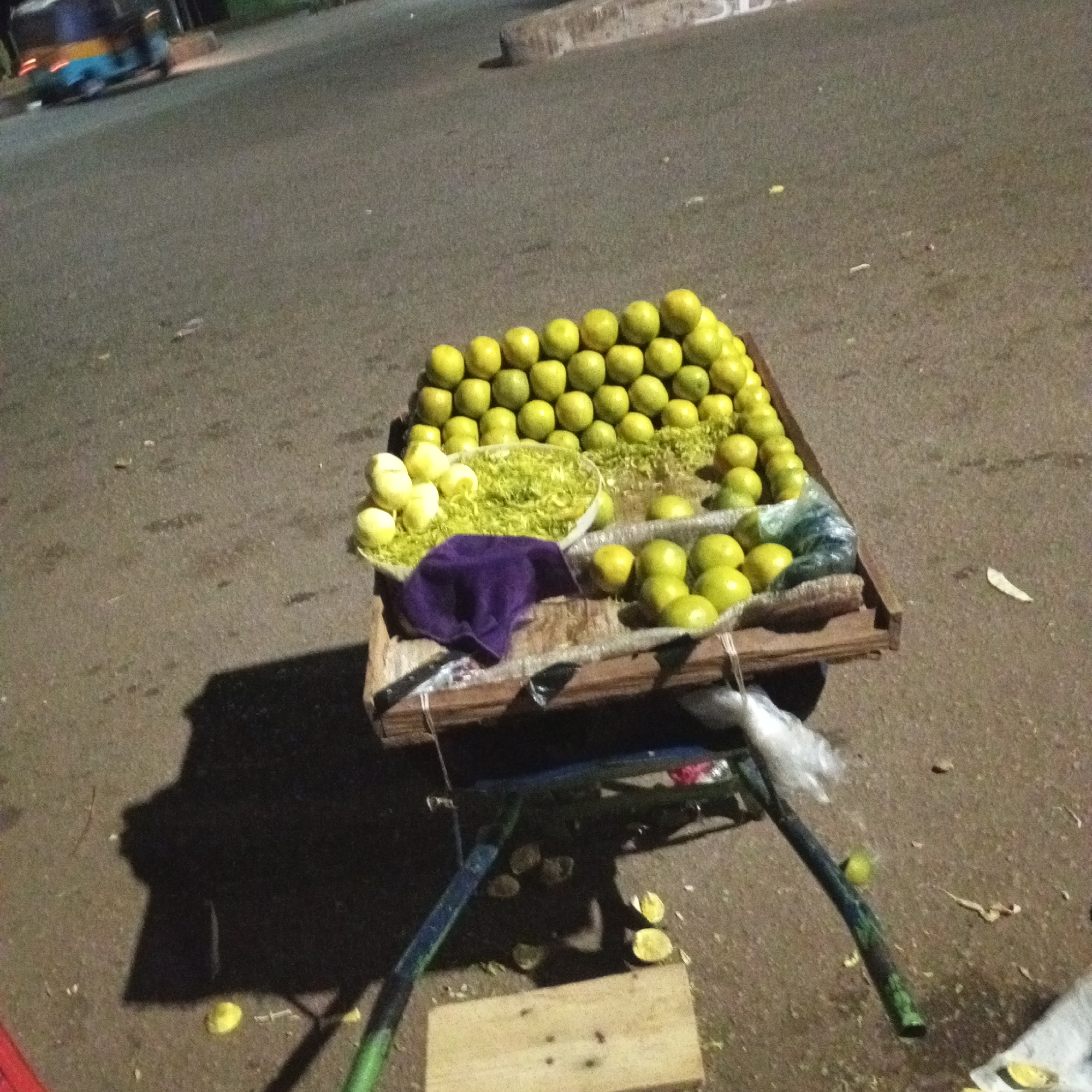 The image depicts a fruit vendor's cart on a paved surface, likely a street in Bukuru, Nigeria. The cart is loaded with lemons, some of which are whole, some peeled, and some shredded in a bowl. A purple cloth sits amidst the lemons. The cart's frame appears to be made of wood and metal, with a single wheel visible. Discarded lemon rinds are scattered on the ground beneath the cart. A portion of a vehicle, possibly a tuk-tuk, is visible in the upper left corner of the image. The scene appears to be captured at night or in low light conditions. There are no people visible in the immediate frame.