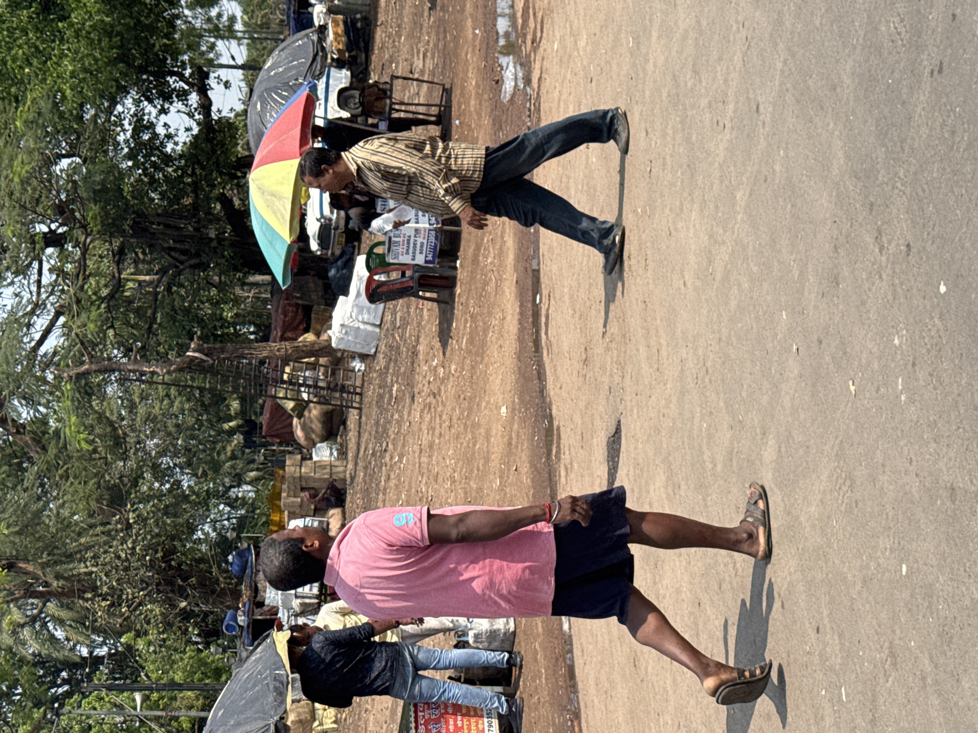 The image depicts a street scene in Kolkata, India. Two men are walking in opposite directions on a paved road. The first man is wearing a pink shirt and dark shorts, while the second man is wearing a striped shirt and blue jeans and carrying a colorful umbrella.
In the background, there is a row of trees on one side and what appears to be a market or street vendors on the other side. There are goods stacked on the ground, along with chairs, and other miscellaneous items. There is an advertising sign present on the street.