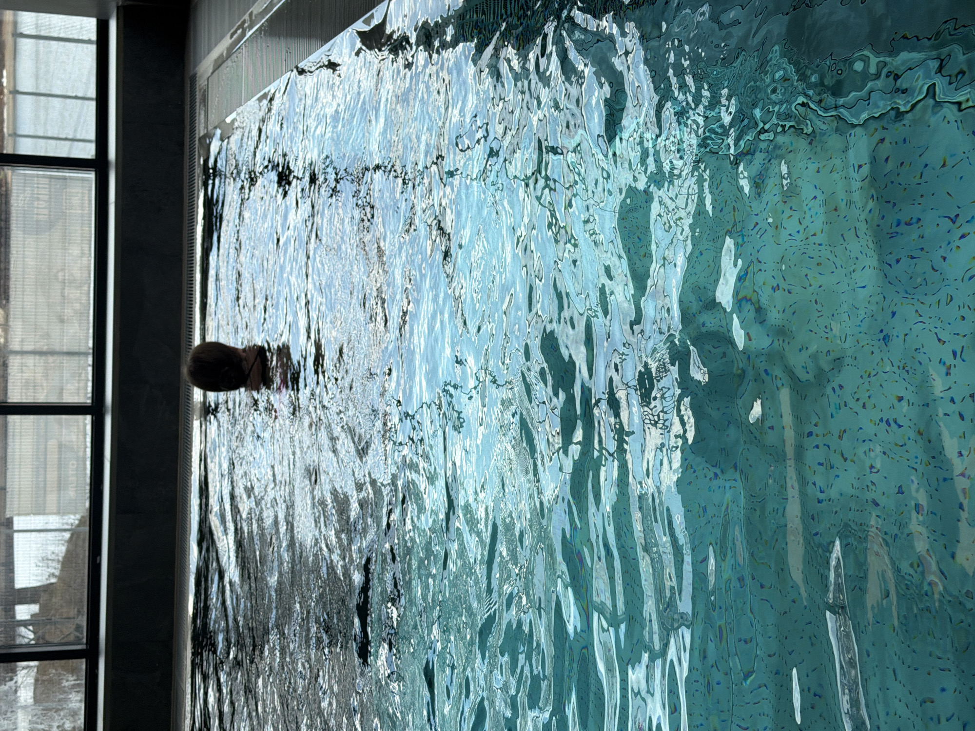 A person is swimming in a pool located inside a building in Chaikovsky, Russia. The pool water is turquoise and rippled. A window with black frames is visible to the left of the pool. The building's interior is modern with dark gray walls. The person's head is visible above the water.