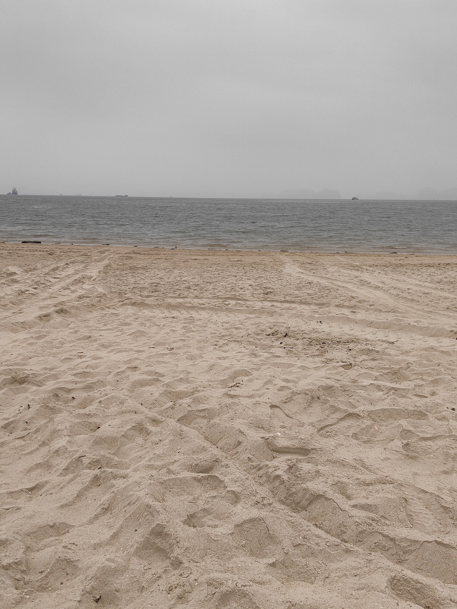 The image depicts a sandy beach in Bai Chay Ward, Vietnam, on a cloudy day. The beach is covered in beige sand with visible tire tracks. In the distance, the sea meets the horizon under an overcast sky. Several cargo ships are sparsely distributed across the horizon line. There are no people visible in the frame.