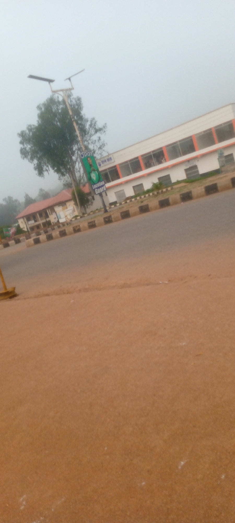 The image depicts a street scene in Gura Top, Nigeria, under an overcast sky. A white and orange building with large windows is visible on the right side of the frame, set back from the road. A tall pole with solar panels stands in front of the building, alongside a green banner that reads "Opay Location Guard" with a portrait of a person and a shield logo. To the left of this, another building with a red roof is partially visible. The foreground is dominated by a dirt road or shoulder. A black and white concrete barrier runs along the edge of the road. The overall atmosphere is hazy and subdued.