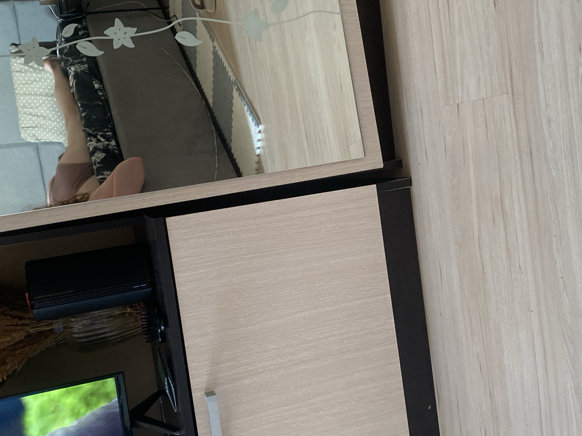 The image shows a fragment of a room interior. In the lower left corner, there's a portion of a TV screen and a black rectangular object mounted on the furniture, possibly a router or media player. The cabinet or furniture piece has a light wood-grain finish door. Above it, there appears to be a mirrored surface with decorative floral designs etched onto it. The reflection in the mirror reveals a sofa or cushion with a patterned cover, and a glimpse of a person's leg and torso. The floor in the right portion of the image has a light wood-like finish. The context doesn't provide sufficient information to identify a specific location in Yekaterinburg, Russia.