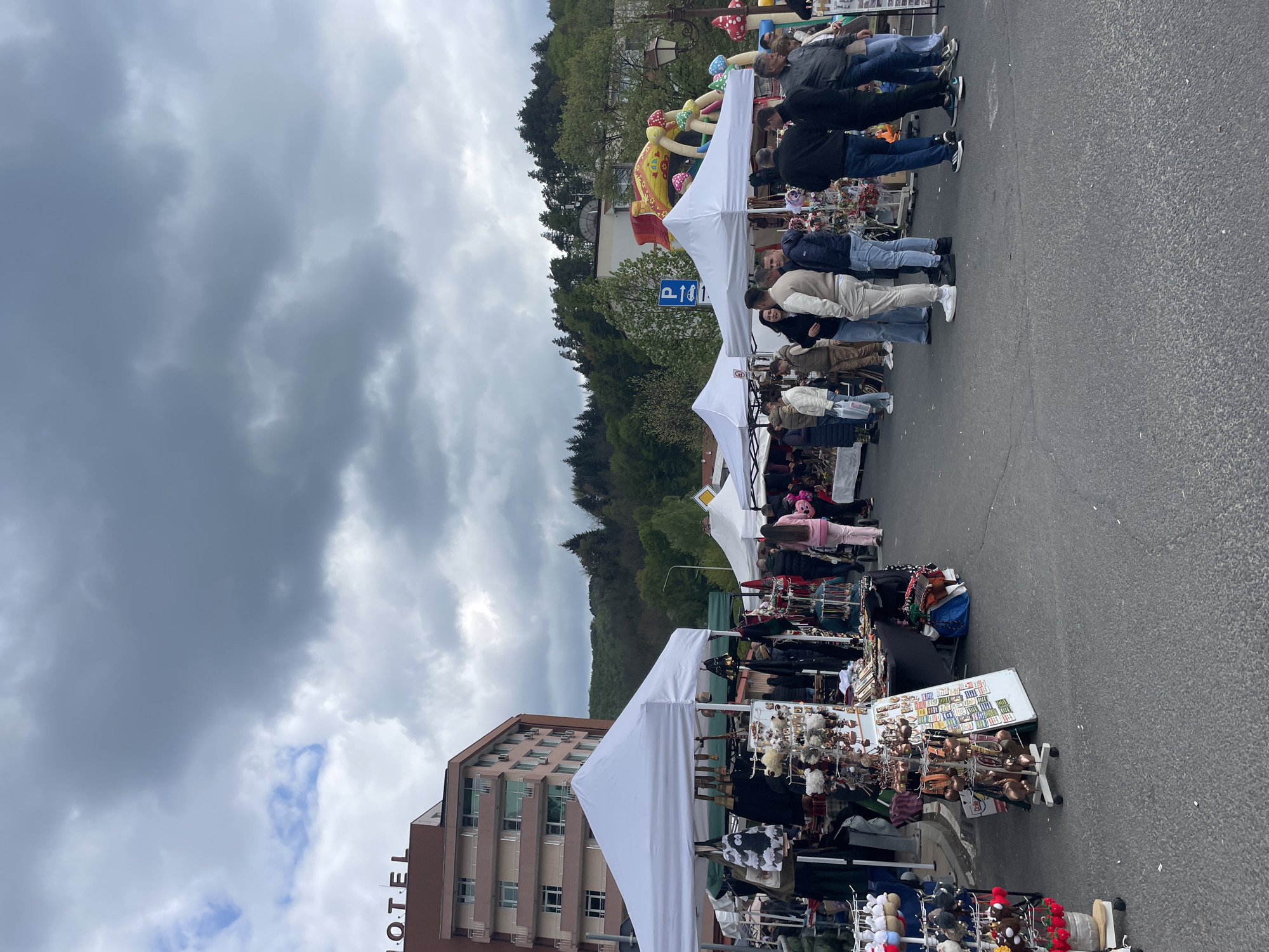 The image shows an outdoor market in Zlatograd, Bulgaria, with white tents set up along a street. People are browsing the stalls, which display various goods for sale. A hotel building is visible on the left side of the frame. In the background are trees and a cloudy sky. There is also a parking sign and a colorful mushroom-themed inflatable structure visible in the distance.
