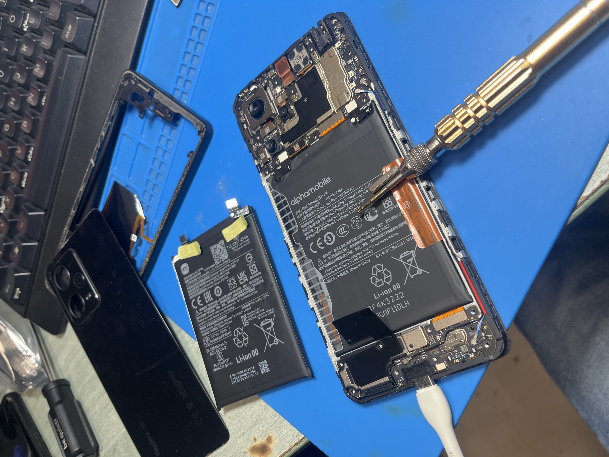 The image depicts a disassembled smartphone on a blue anti-static mat, likely during a repair process. The smartphone is an "alphamobile" brand, model BP4K. A second, identical battery is beside the disassembled phone. The phone's back cover, bearing the brand "Redmi", is also visible. A screwdriver is positioned near the phone's battery, and a USB cable is plugged into the phone. A keyboard is partially visible on the left side of the frame. A black screwdriver labeled "be quiet!" is also partially visible at the bottom left corner.