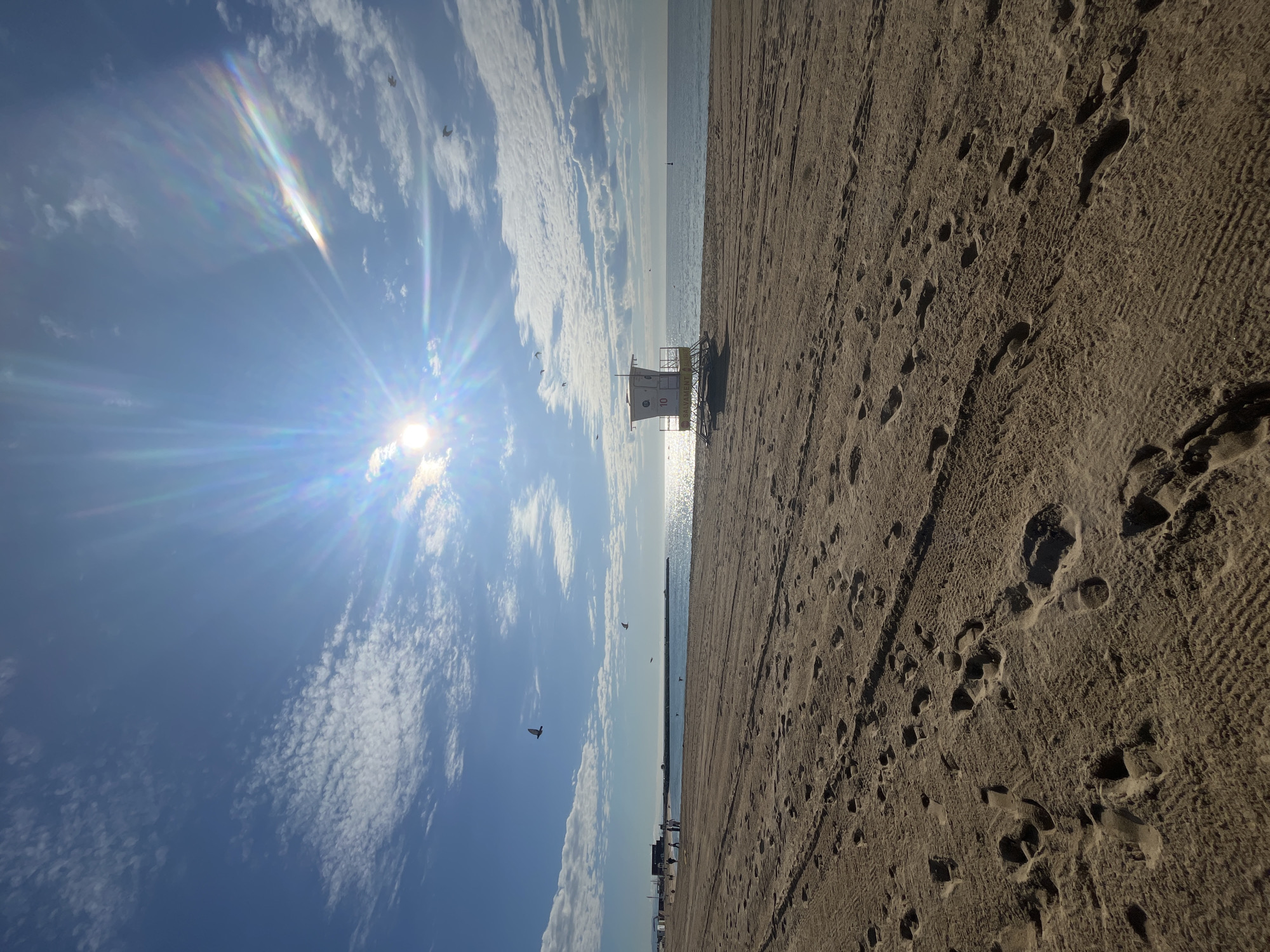 The image shows a beach scene, likely taken in Barcelona, Spain, given the typical lifeguard tower style. The sand is light brown and covered with numerous footprints. A white lifeguard tower stands near the shoreline, casting a shadow on the wet sand. The sun is bright, creating lens flare and reflecting off the water. The sky is blue with scattered white clouds. Several birds are flying in the sky. A pier is visible extending into the water. Some sunbathers are visible in the distance.
