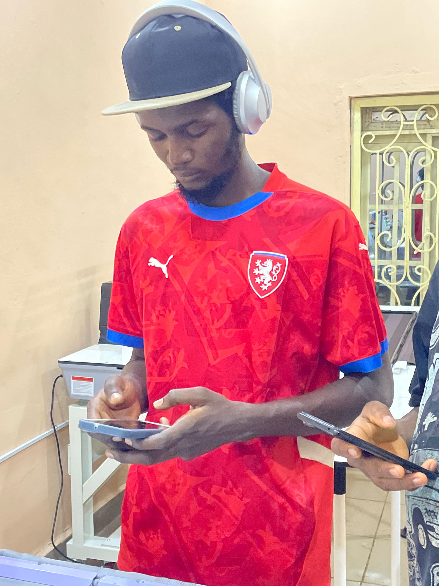 In Gura Top, Nigeria, a young Black man wearing a red Puma soccer jersey with a blue collar and cuffs, a black baseball cap, and white headphones, is focused on his smartphone. He holds the phone in his left hand while his right hand rests on it. Another person's hand holding a black smartphone is visible to the right of the frame. The background features a plain, light-colored wall and a metal-framed window with decorative grilles. A white stand supporting a scale is also visible to the left.