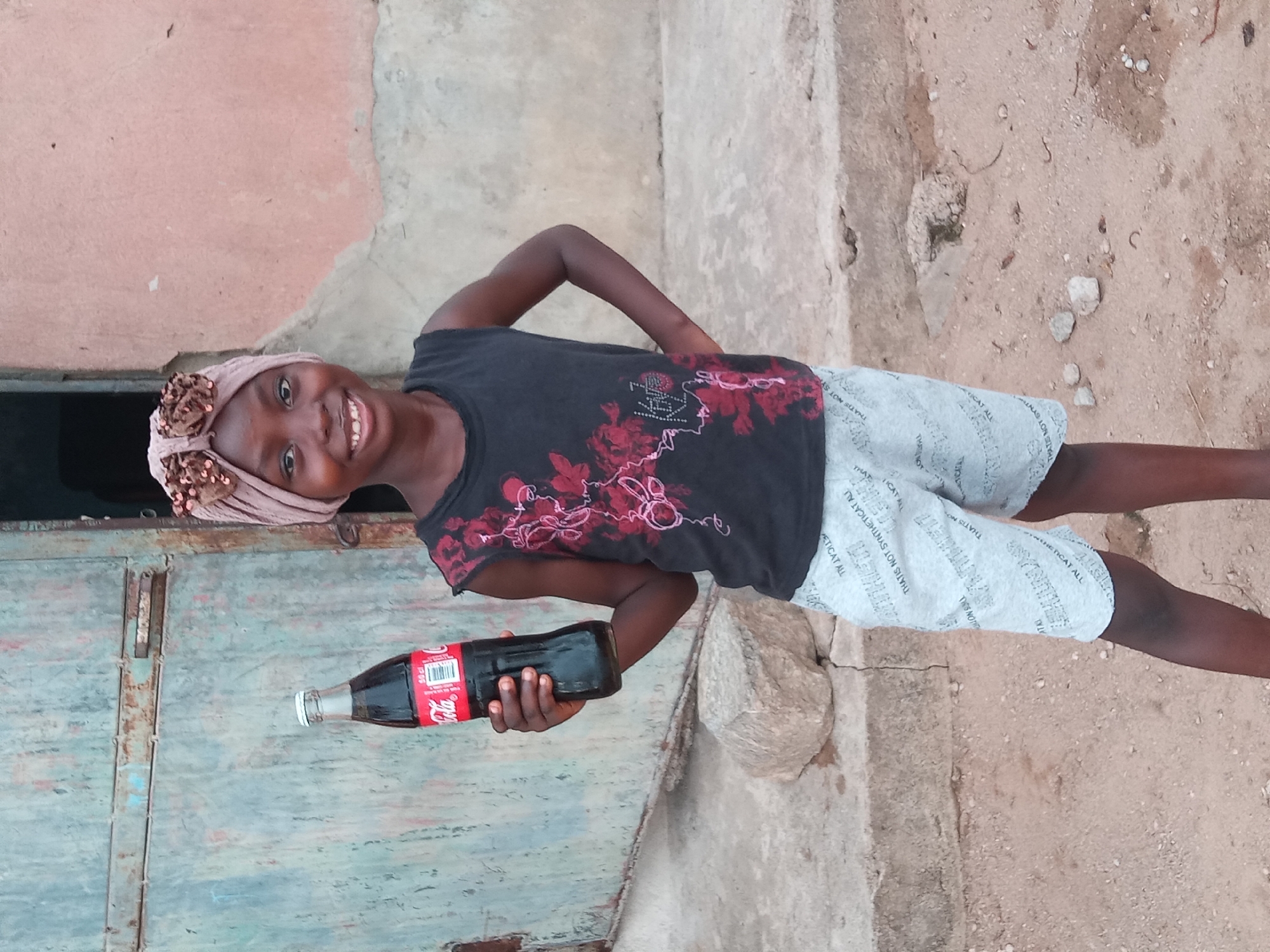 A young girl stands in front of a weathered, turquoise-painted metal door in Sabon Tasha, Nigeria. She wears a dark, sleeveless shirt with a red floral design, light-colored shorts printed with text, and a decorated headwrap. She holds a glass Coca-Cola bottle in her left hand, smiling at the camera. The background includes a crumbling pink wall and a dirt ground strewn with rocks and debris.