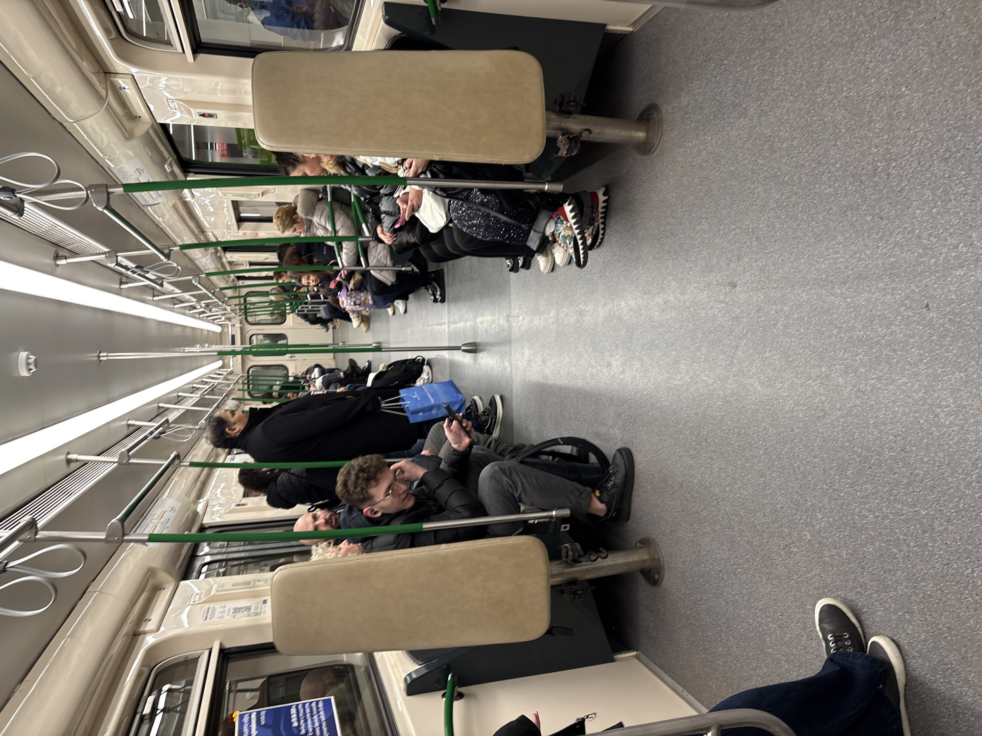The image captures the interior of a metro train in Sofia, Bulgaria. The carriage is populated with several passengers seated and facing the direction of travel, others are standing. Most appear to be occupied with their own devices, or in quiet contemplation. The train's interior features light-colored walls and seats, with green handrails running along the length of the carriage. A pair of feet wearing black and white sneakers are visible in the lower right corner, suggesting the perspective of an unseen passenger or an observer. A display showing transportation information is seen next to a door on the left. The floor of the train car is a speckled grey.