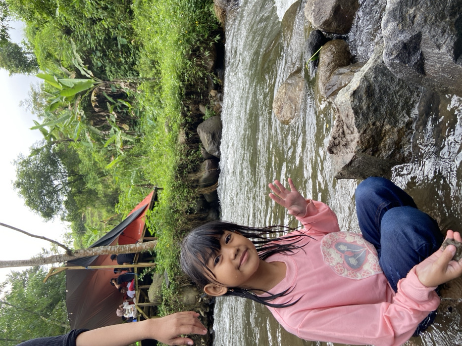 A young girl with wet hair and bangs sits in a flowing stream among large rocks, wearing jeans and a long-sleeved pink shirt with a floral patch. She smiles and raises her hand in a wave. In the background, lush green vegetation covers the landscape. A tent-like structure with people gathered under it is partially visible. The stream rushes past, creating a scenic and refreshing outdoor environment.