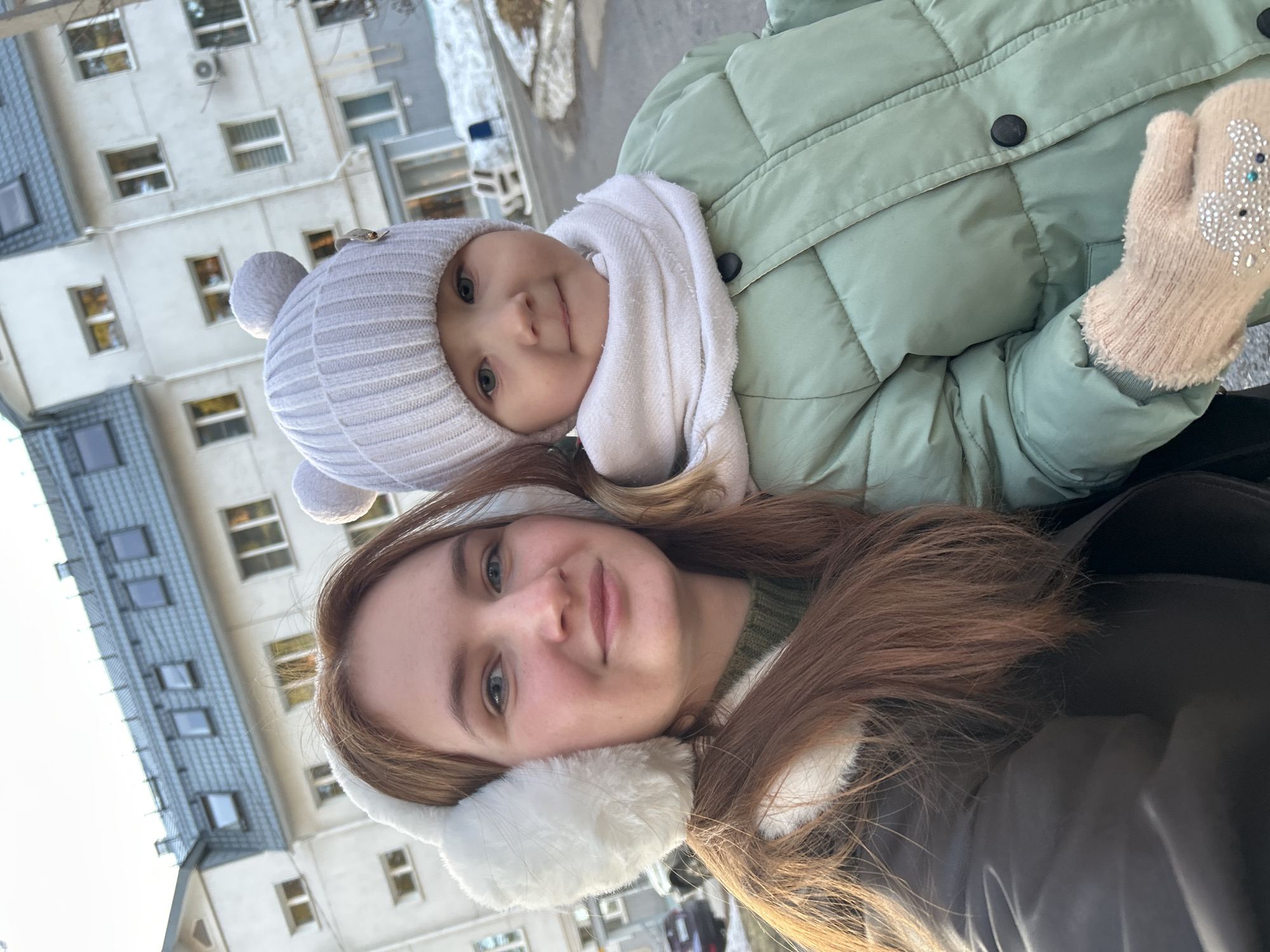 A woman and a baby are in the frame. The woman has light skin, long brown hair, and blue eyes, and she is wearing white earmuffs. The baby is fair-skinned and is wearing a gray hat with pom-poms, a white scarf, and a light green quilted jacket. The baby is also wearing a light beige mitten with some embellishments. They are outdoors in front of a building, possibly an apartment complex, with many windows. The photo appears to have been taken in daylight, with the sun casting shadows. Based on the architecture of the buildings and information from online sources, this photograph was likely taken in the city of Chaikovsky, Russia.