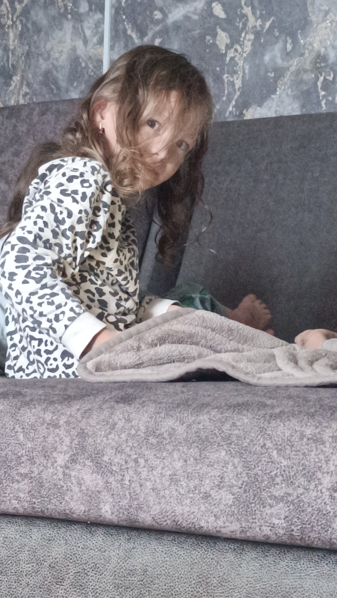 The image features a young girl with long, dark, curly hair that partially obscures her face. She is wearing a black and white leopard-print shirt with white cuffs. She is sitting on a gray sofa, leaning towards the camera and looking directly at it. Her body is angled away from the camera, with her left arm bent and resting on the sofa. A gray blanket is draped over the sofa in front of her. To her right, her legs are visible, with her feet peeking out from under the blanket. Another person's bare foot is visible to the right of the girl's feet, suggesting another person is also on the sofa. The background appears to be a wall with a textured, marble-like pattern. The lighting suggests it is daytime, and the indoor setting indicates a home environment. The girl's expression is somewhat ambiguous due to her hair covering part of her face. The scene conveys a casual, relaxed atmosphere.