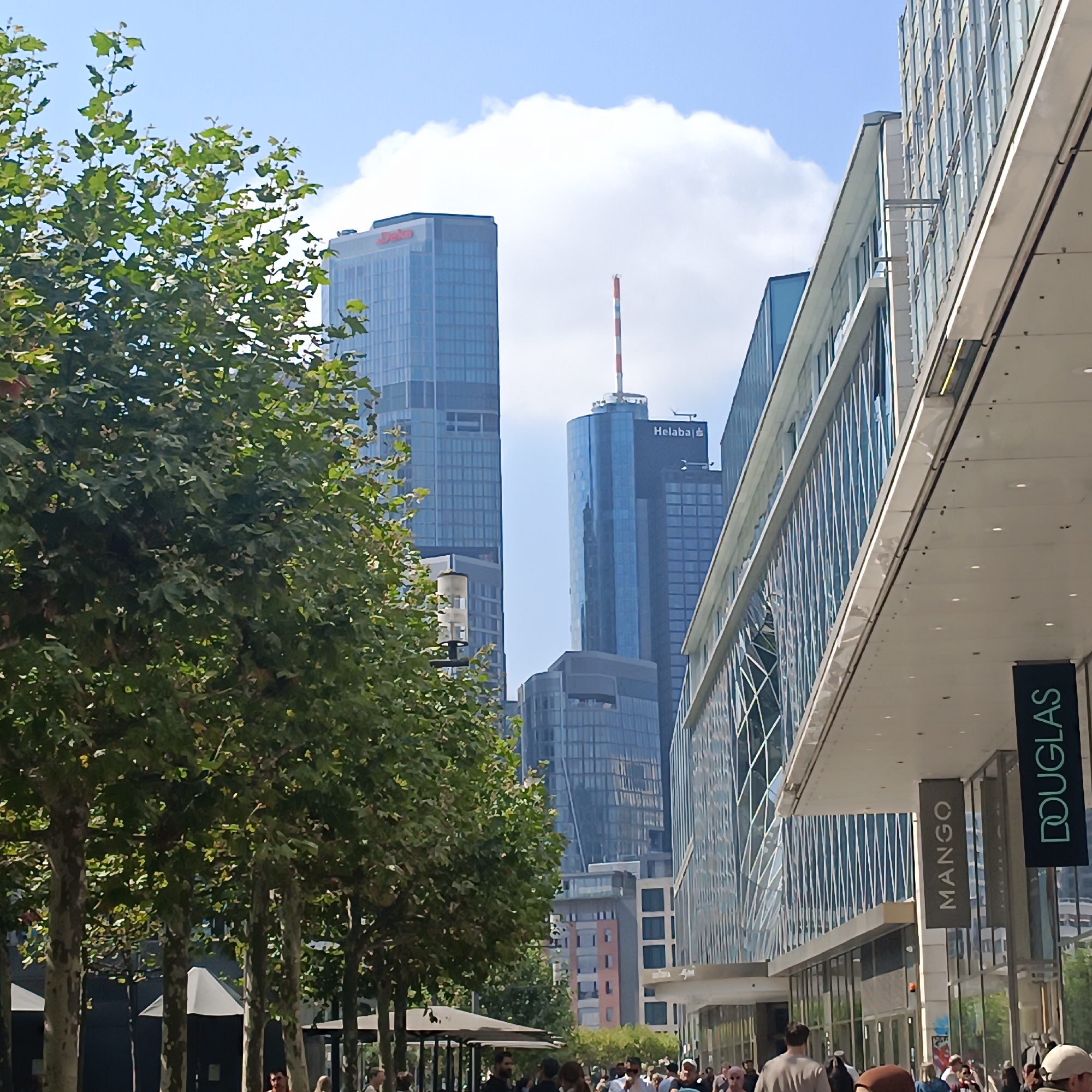 In Frankfurt, Germany, on a bright, sunny day, a street view captures a line of trees in the foreground, their leaves casting dappled shadows. Beyond the trees, modern skyscrapers dominate the background. To the left, a tall, blue-glass skyscraper with a red logo, possibly "Deka," stands prominently. To the right, another sleek, glass-fronted building features prominent signage for "MANGO" and "DOUGLAS." A crowd of people walks along the street, suggesting a commercial or public area within the city. The architecture indicates a contemporary urban setting, characteristic of Frankfurt's financial district.