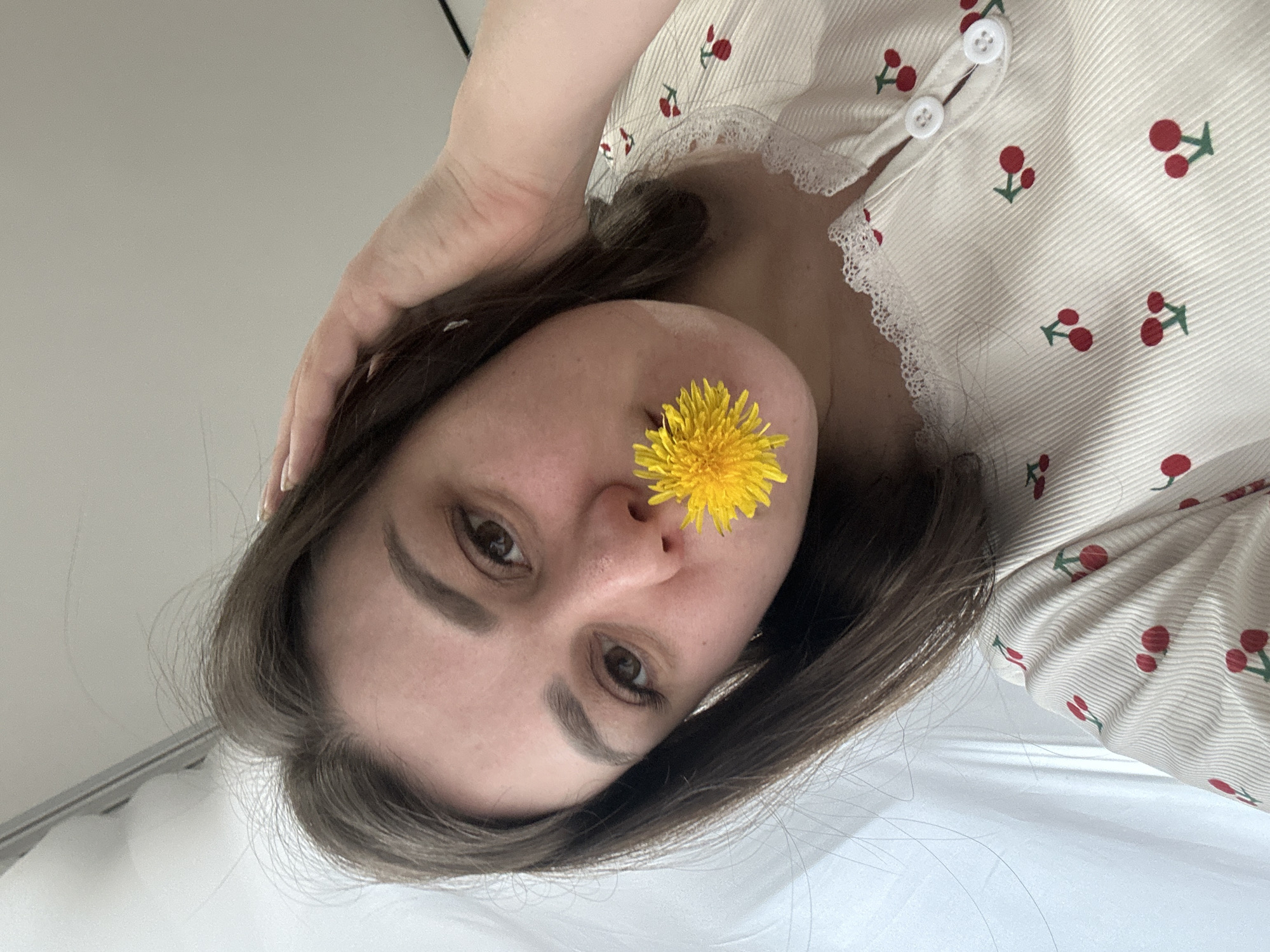 A woman is lying down, holding a yellow dandelion flower against her nose. She is wearing a light-colored pajama top with red cherry patterns and white lace trim. She has brown hair and brown eyes. The background is blurred, showing a white surface. The image appears to have been taken in Chaikovsky, Russia.