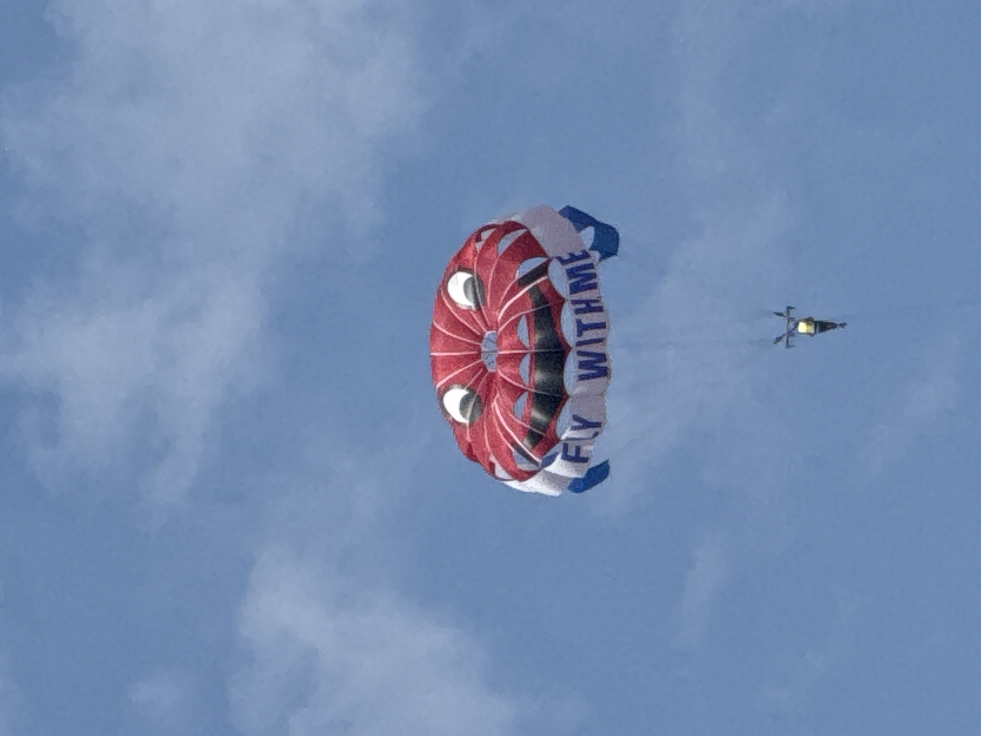 A red, white, and blue parachute designed to look like a smiling face with large white eyes and black pupils is airborne against a clear blue sky with scattered white clouds. The parachute has the words "FLY WITH ME" printed on its white underside. A person is suspended beneath the parachute by a harness and tow rope. The scene is observed from below, looking up at the sky. The image does not contain any visual cues indicating the city of Boreti, Montenegro.