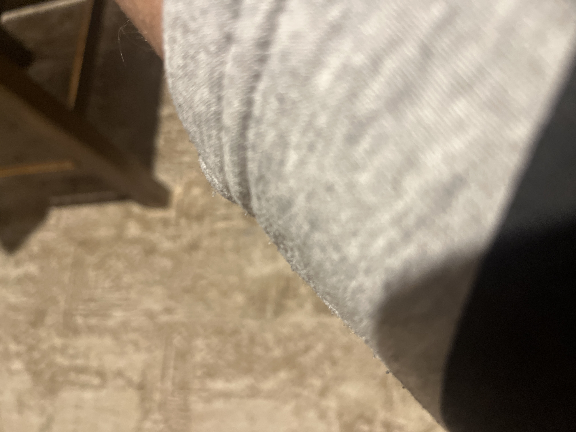 A close-up, low-angle, and out-of-focus indoor image depicts a scene in Blagoevgrad, Bulgaria. The immediate foreground features a section of light grey, possibly ribbed or knit fabric, likely a garment, with a dark, indistinct shadow or object obscuring its lower left portion. To the upper right of the fabric, a patch of human skin with visible fine hairs is present, suggesting an arm or leg. The midground and background consist of a blurry, light beige to brown textured surface, consistent with a carpet or rug, exhibiting subtle patterned impressions. Further in the upper right background, out-of-focus dark brown angular structures, indicative of wooden furniture such as chair or table legs, are partially visible.