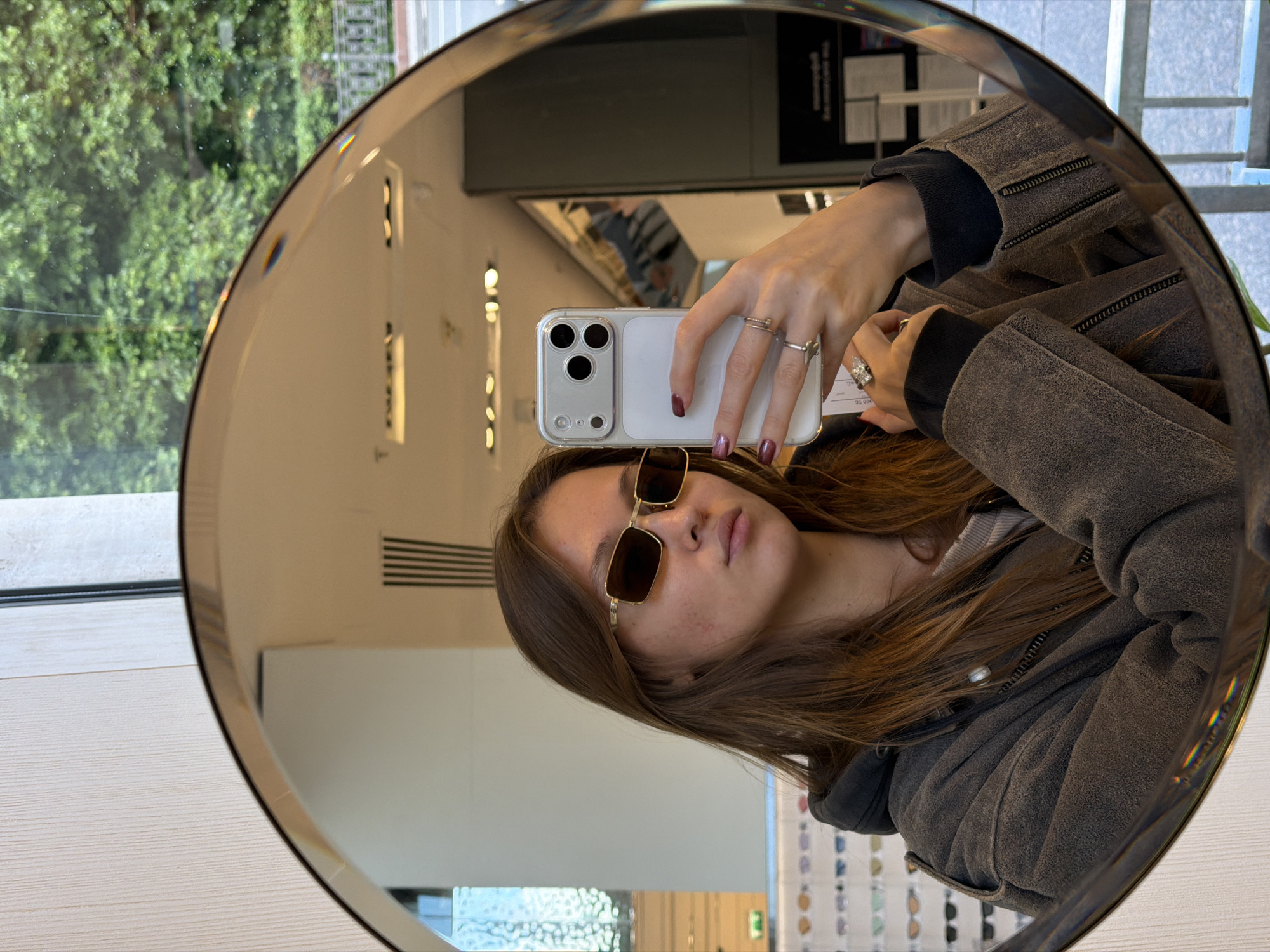 A woman is taking a selfie in a round mirror, presumably inside a store. She is wearing sunglasses and a jacket. In the reflection, eyeglasses are visible at the bottom, and trees can be seen outside the window on the left. The location may be Moscow, Russia.