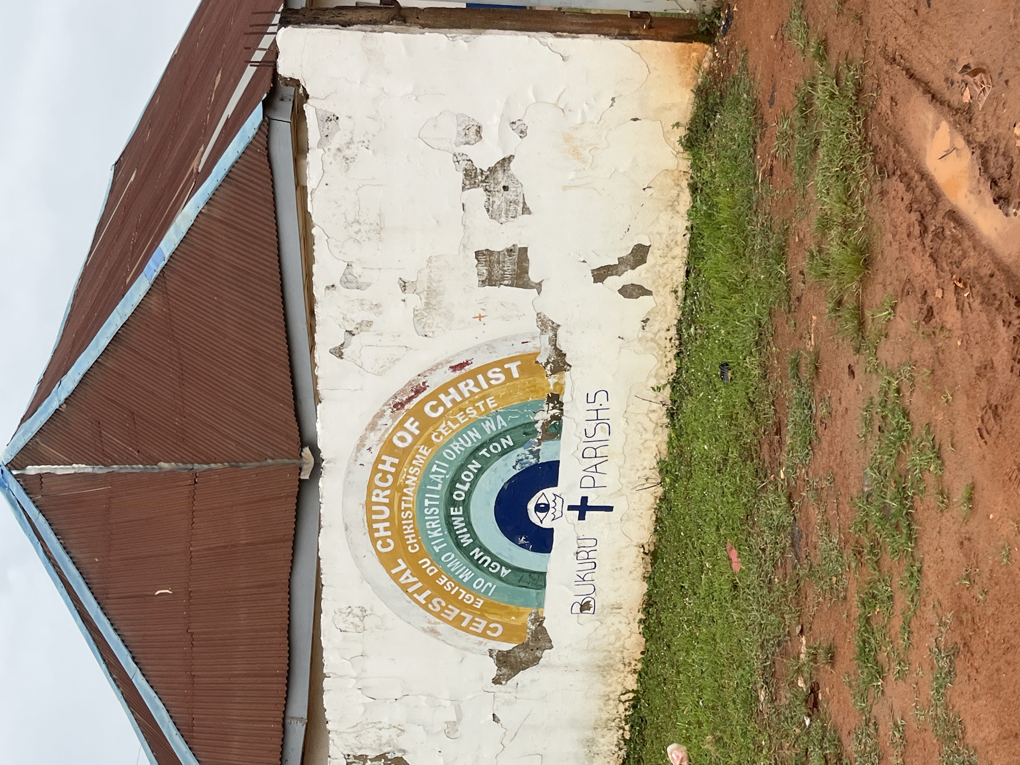 The image shows a weathered white wall with a mural depicting a rainbow and text. The mural is centered on the wall, above a patch of overgrown grass and a muddy dirt area. The text in the mural reads "CELESTIAL CHURCH OF CHRIST" in a semi-circle. Inside the rainbow, there are concentric circles of color with additional text. At the bottom of the mural, the words "BUKURU PARISH" are written, with a cross and an eye symbol. The wall itself shows signs of age and damage, with peeling paint and cracks. The background features a rust-colored corrugated metal roof with blue trim. The environment appears to be outdoors, likely in a rural or semi-rural setting, given the unpaved ground and vegetation. The sky is overcast. There are no people visible in the image. The location is identified as Bukuru, Nigeria, as indicated by the text on the mural.