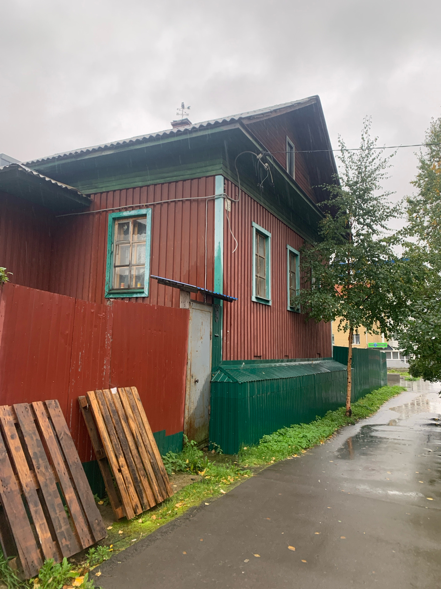 The image depicts a scene on a cloudy, rainy day in Arkhangelsk, Russia. The foreground features a wet asphalt path running alongside a vibrant green metal fence. To the left of the path, leaning against a dark red corrugated metal fence, are two wooden pallets. Behind the green fence, a weathered, dark red wooden house with green trim is visible. The house has at least three windows with light blue or green frames and a small, gray metal door. A young birch tree with green leaves stands beside the path, with more trees and buildings in the background. The sky is overcast, and rain appears to be falling, as evidenced by the wet surfaces and streaks on the house.