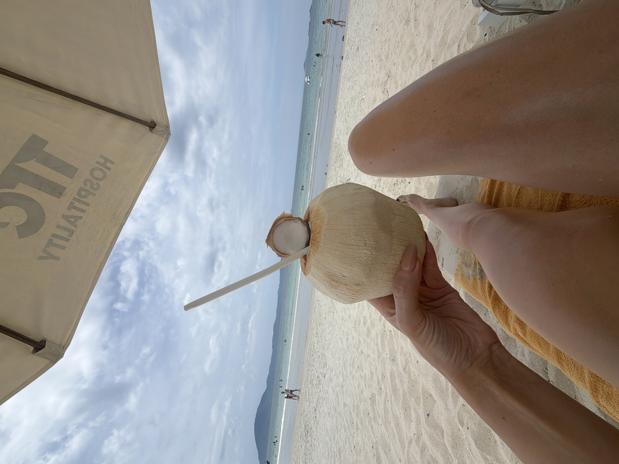 In Dong Ninh Hoa Ward, Vietnam, a first-person perspective shot captures a tranquil beach scene. A person's hand holds a freshly opened coconut with a straw, the white flesh and liquid visible. The coconut is positioned in the foreground, with a sandy beach stretching out behind it. In the middle ground, people are scattered along the shore and in the shallow, turquoise water, some wading and others relaxing. The background reveals a hazy coastline with mountains under a cloudy, overcast sky. An umbrella, partially visible at the top of the frame, provides shade. The overall ambiance is one of relaxed leisure on a tropical beach.