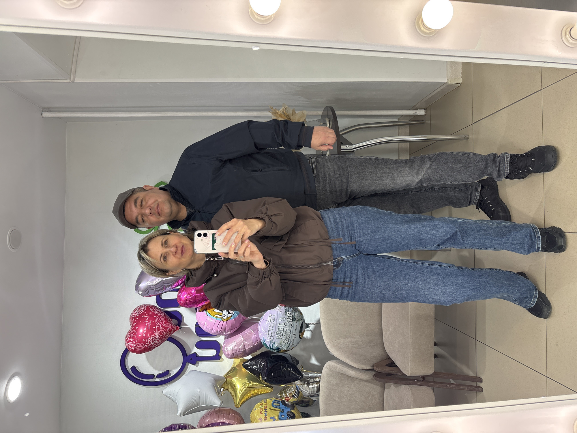 A middle-aged man and woman are posing for a selfie in a mirror. The woman is holding the smartphone. Behind them, there are several colorful balloons. In the background, a logo with the silhouette of a woman. According to the balloons, the location is in the city of Nizhnevartovsk, Russia. The man is wearing a baseball cap, a dark jacket, and jeans. The woman is wearing a brown jacket and blue jeans. There is a clothing rack visible as well as chairs. The reflection is from a mirror with lightbulbs around the edges. The floor appears to be tan tile.