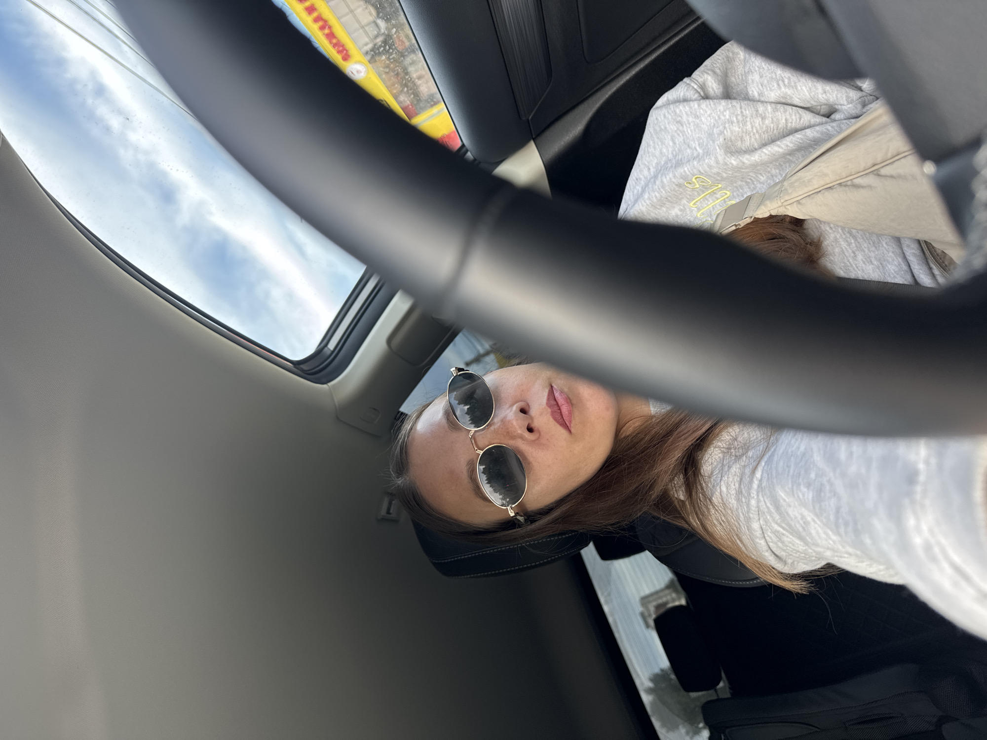 A young woman is sitting in the driver's seat of a car. She is wearing a grey sweatshirt and oval-shaped sunglasses. The car has a sunroof open, showing a cloudy blue sky. In the background, through the car window, there is a yellow building with Russian text, suggesting the location is in Chaikovsky, Russia. The steering wheel is prominently in the foreground, partially obscuring the woman and the interior of the car.