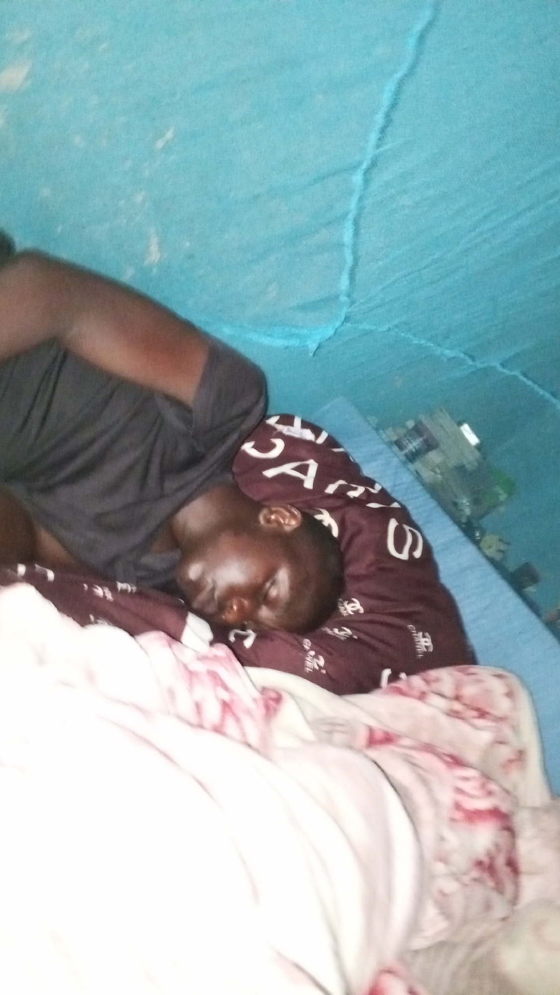 A person is sleeping in a bed under a blue mosquito net. The bed has a blue mattress, a dark brown and white patterned pillow, and a white and pink patterned blanket. The person is wearing a black t-shirt. Several blurry objects are in the background near the head of the bed. The location and city are unknown.