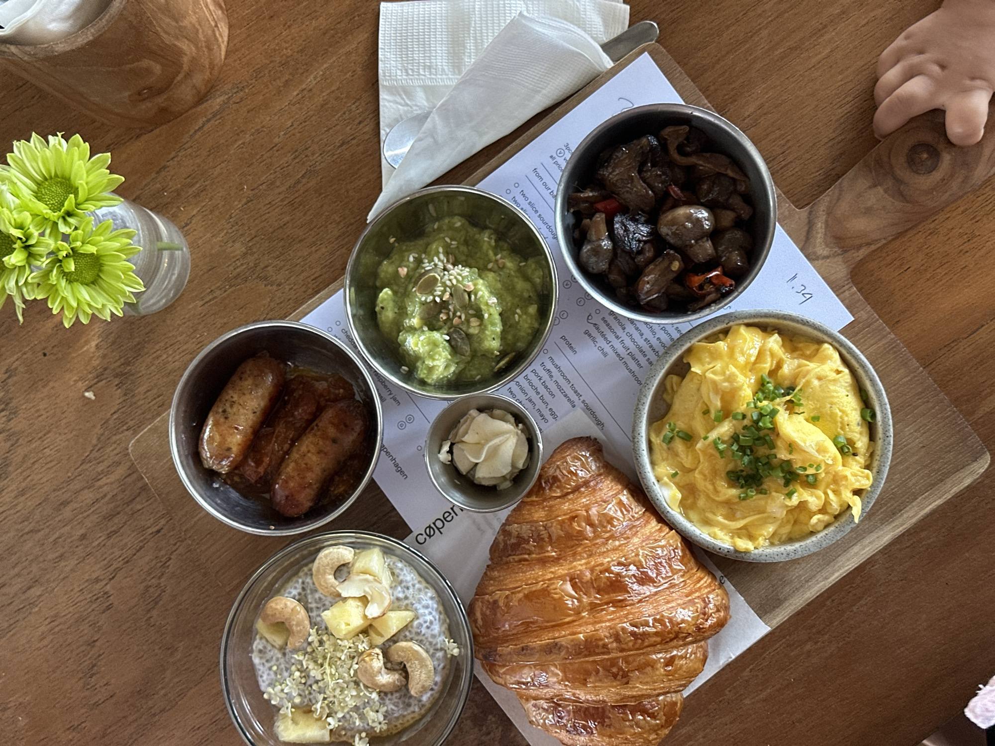 An overhead, eye-level shot captures a breakfast spread on a wooden table. The spread includes a flaky croissant, a bowl of scrambled eggs with chives, a small dish of pickled ginger, a bowl of avocado with pumpkin seeds and sesame seeds, a bowl of dark sautéed mushrooms with chili peppers, and a glass bowl of chia seed pudding with pineapple chunks and cashews. Three breakfast sausages are in a small metal bowl. A white napkin with a spoon is to the right of the avocado bowl. A small vase with green chrysanthemums is to the top right. A child's hand is visible at the bottom right, reaching for the wooden serving board. The wooden table has a visible grain. The scene is located in North Kuta, Indonesia.