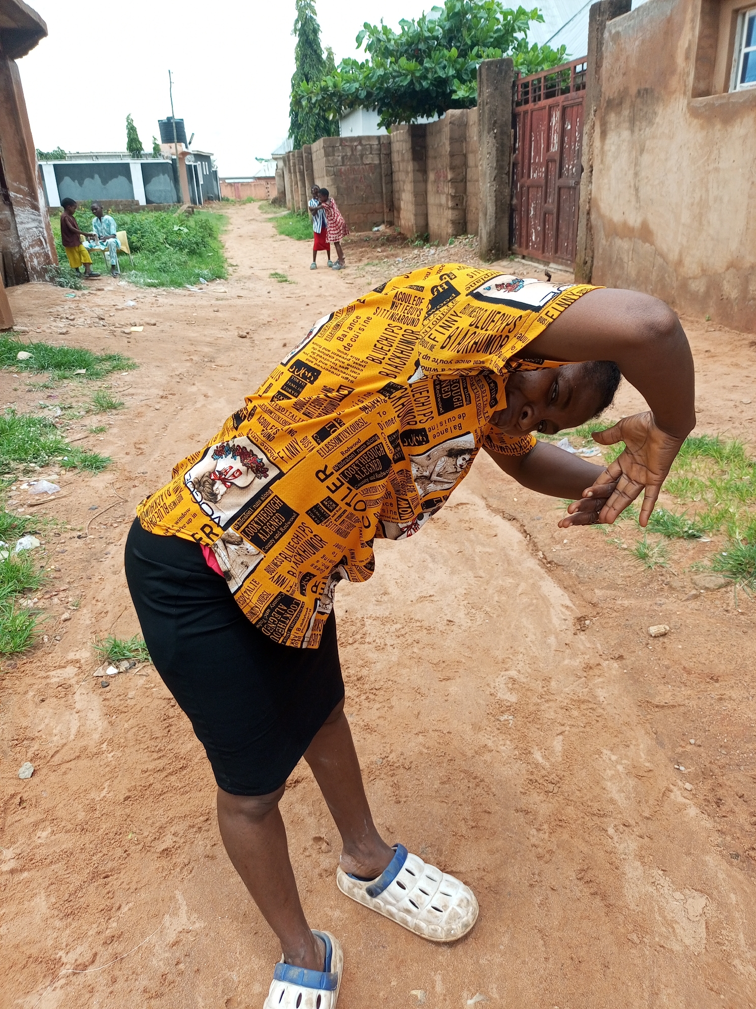 In Tempure, Nigeria, a young woman is captured from the waist down, bending over with her head tilted downwards. She is wearing a black skirt and a vibrant yellow t-shirt adorned with a repetitive pattern of text and images. Her arms are bent at the elbows, with her hands open, palms facing away from her body. She is wearing white clogs with blue trim. The ground is composed of reddish-brown dirt, with patches of dry grass growing sporadically. In the background, a pathway stretches towards a few buildings. To the left of the pathway, there is a cluster of green vegetation and a water tank atop a structure. Further in the background, two young boys can be seen standing near some buildings, one in a brown shirt and yellow shorts, the other in a light blue shirt and patterned pants, and two young girls in a red and white dress embrace each other. The overall scene suggests an outdoor setting in a Nigerian community.