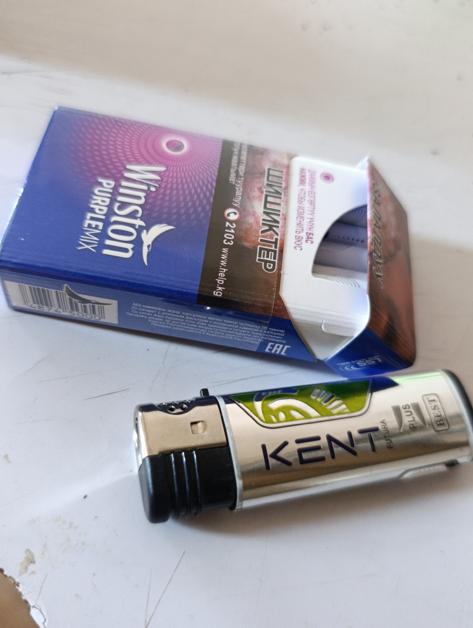 The image shows a Winston Purple Mix cigarette pack and a Kent lighter on a white surface. The cigarette pack is partially open, revealing the cigarettes inside. The Kent lighter is silver with green accents and has the brand name "KENT" printed on it, along with "FUTURA," "PLUS," and "BEST." The background is a plain white surface, likely a table or floor. There are no people visible in the image. The objects suggest a casual setting. No specific details about the city of Tokmok, Kyrgyzstan, are visible in the image itself.
