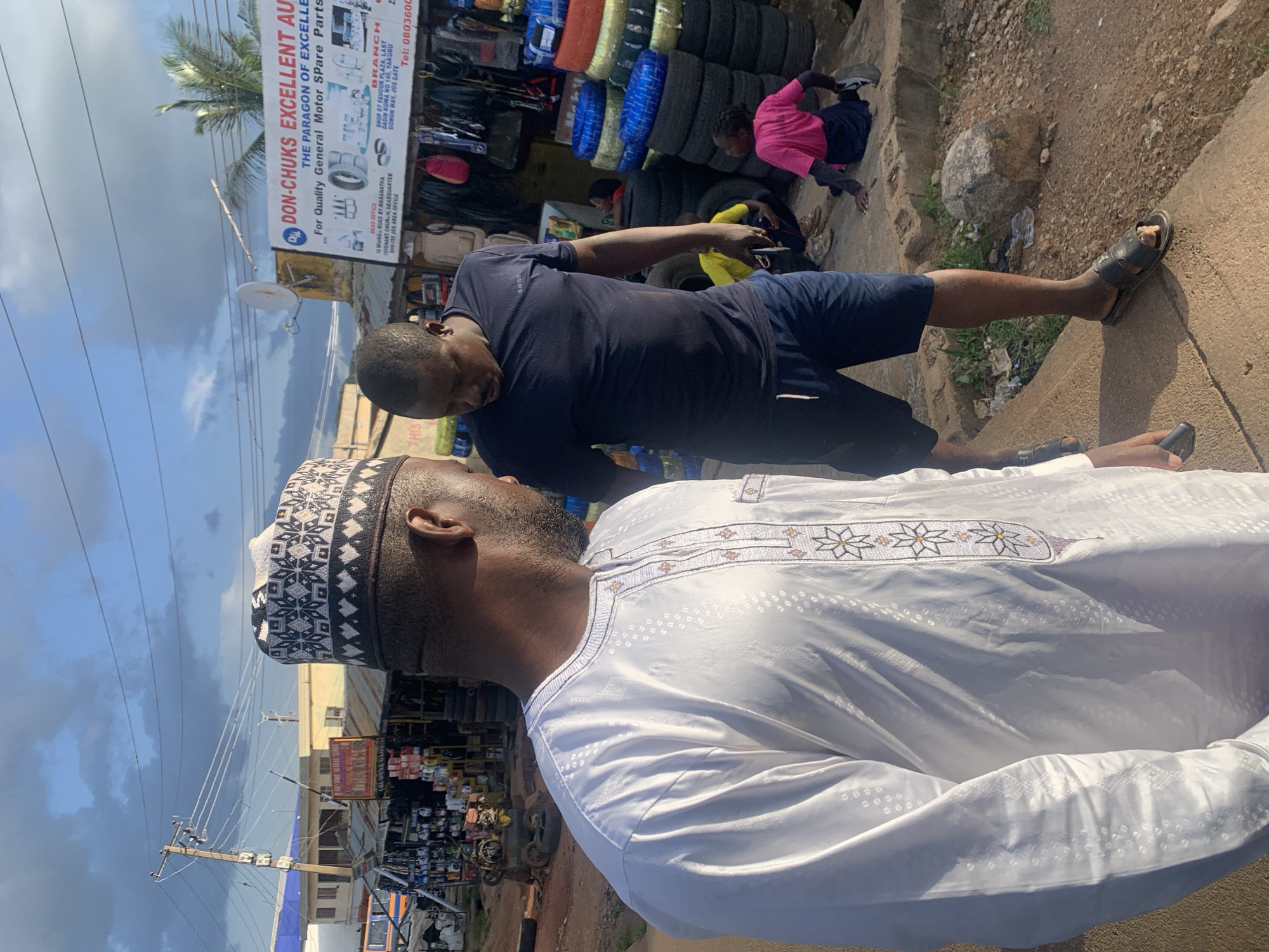 This media file depicts an outdoor scene in Jos, Nigeria. Two men are the primary subjects. In the foreground, a man wearing a white traditional outfit with intricate embroidery and a patterned cap is seen from the shoulder up, facing away from the camera and looking towards another man. The second man, wearing a dark blue t-shirt and shorts, is also facing away and appears to be interacting with the first man or looking at something off-camera.
Behind them, a busy street-side market is visible. Stacks of tires are prominent, suggesting a tire shop or a general auto parts store. A sign for "DON-CHUK'S EXCELLENT AUTO" with text detailing "THE PARAGON OF EXCELLENCE" and "For Quality General Motor Spare Parts" is prominently displayed, along with branch and head office addresses and a phone number. Other market stalls with various goods can be seen in the background, along with people, including a child wearing a pink shirt and dark shorts.
The setting appears to be during daylight, with shadows suggesting it might be mid-morning or late afternoon. The sky is partly cloudy. The overall impression is that of a bustling commercial area.