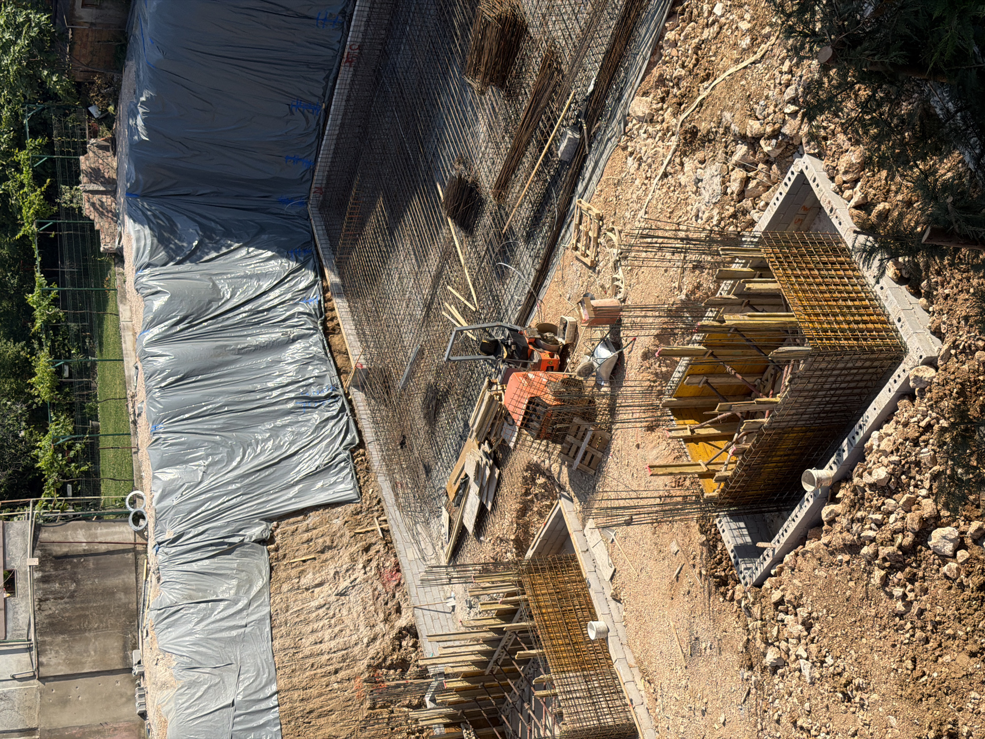 The image depicts a construction site in Bar, Montenegro. A large area is covered with gray plastic sheeting. Adjacent to the sheeting is a partially constructed concrete foundation with extensive rebar reinforcement. The foundation includes a rectangular section filled with wooden forms and more rebar. An orange concrete vibrator sits near the rebar. The surrounding ground is covered with dirt, gravel, and some vegetation.