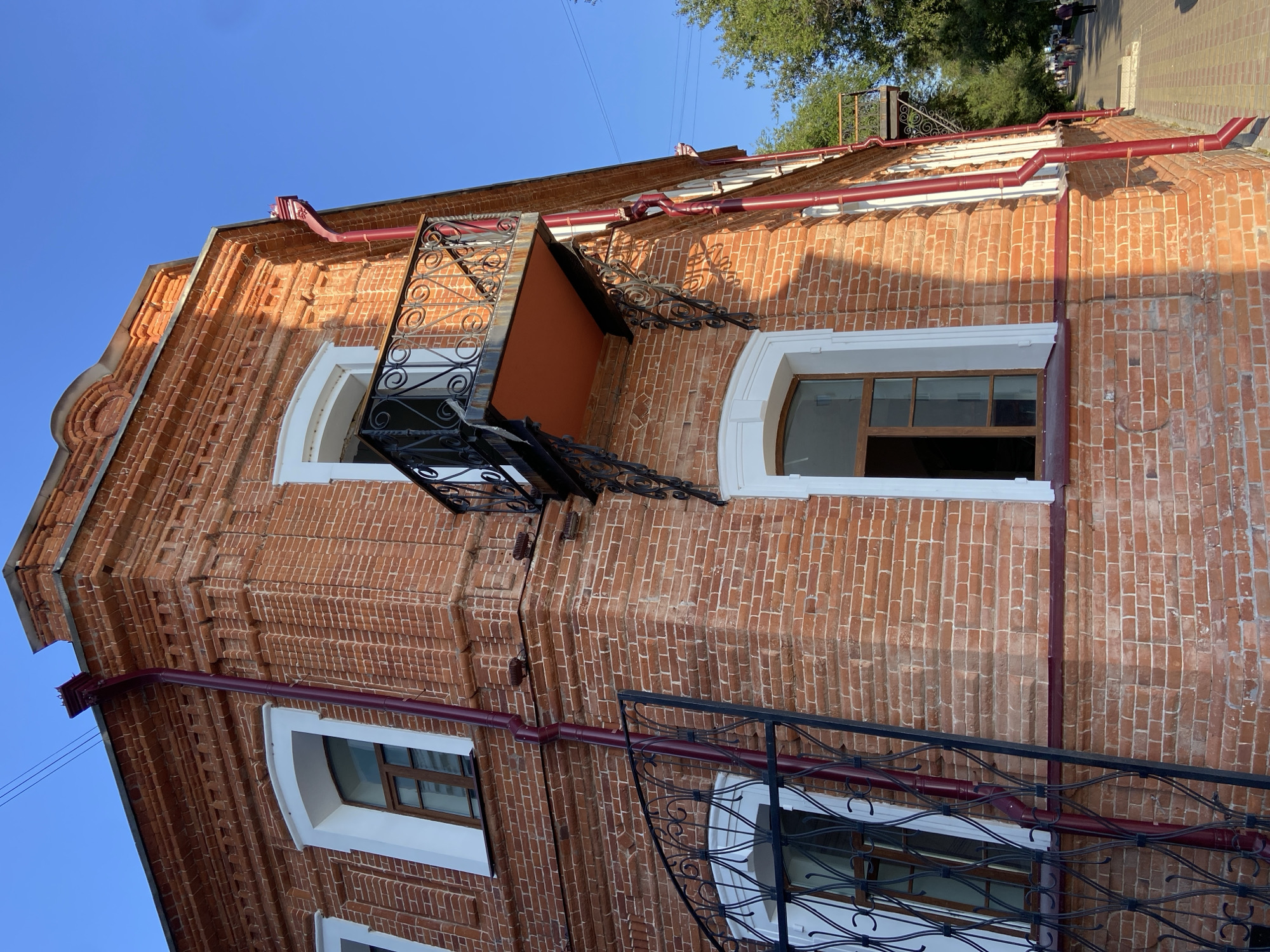 The image shows a corner view of a brick building in Blagoveshchensk, Russia, under a clear blue sky. The building's facade is constructed of red brick, featuring arched windows with white trim and brown wooden frames. A small balcony with ornate black metal railings is attached to the upper story. Red downspouts run vertically along the brickwork. In the background, a tree-lined sidewalk with a few pedestrians suggests an urban setting. The lighting indicates it is daytime.