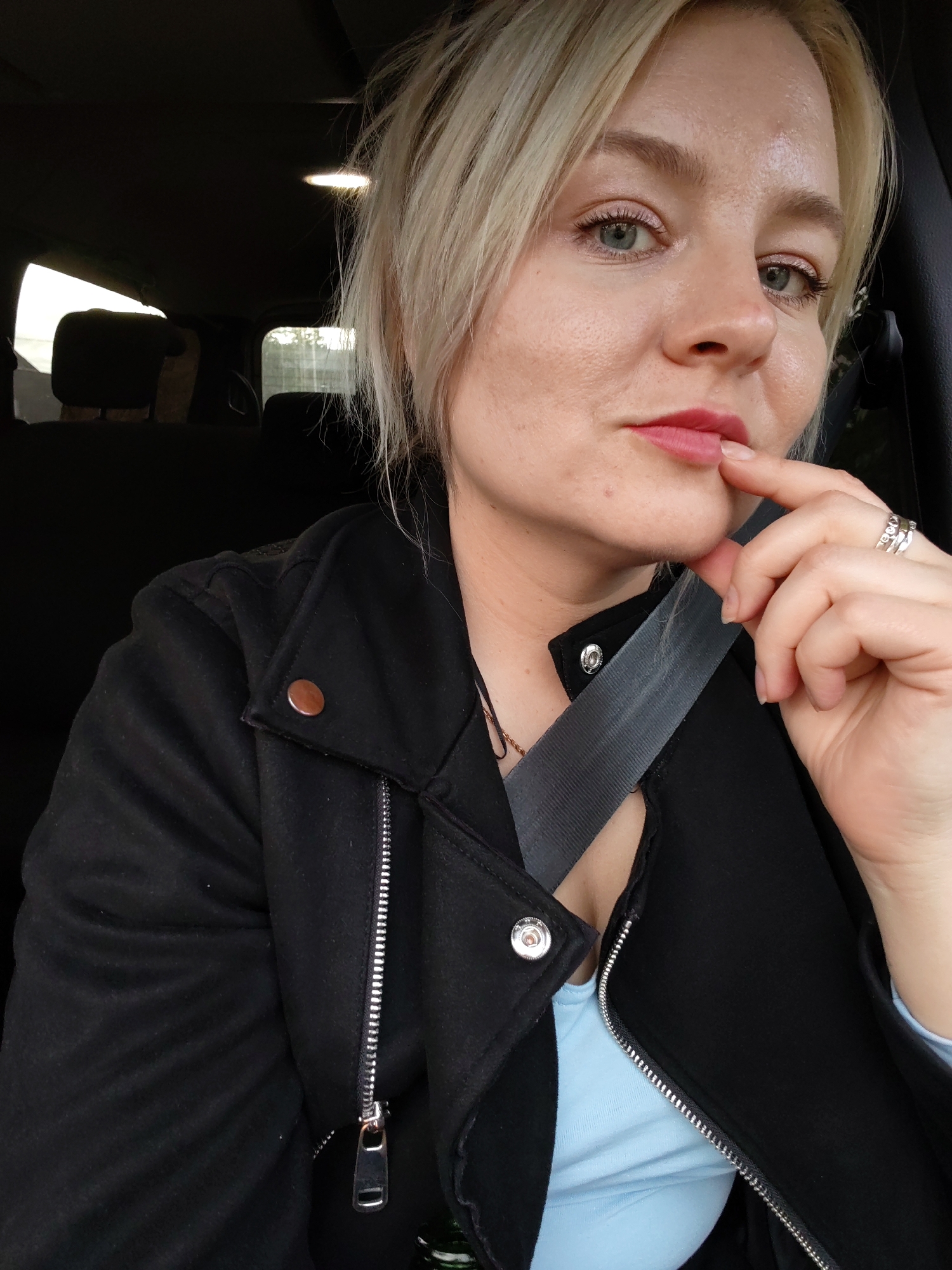 The image displays a close-up, head-and-shoulders portrait of an adult female seated inside a vehicle. She has short, slightly disheveled blonde hair and light-colored eyes, looking directly at the camera. Her lips are painted a soft red-pink, and her right index finger is positioned against her lower lip. She wears several silver-toned rings on her left ring finger. The woman is dressed in a black jacket featuring a prominent silver zipper and a round, bronze-colored stud on the lapel, along with a silver snap button visible near the collar. A light blue top is visible beneath the unzipped section of the jacket. A grey seatbelt diagonally crosses her chest. The background shows the dark interior of the car, including the tops of rear seats and diffused natural light visible through the windows, suggesting an outdoor environment with some foliage. An interior light fixture is visible on the dark overhead panel of the vehicle.