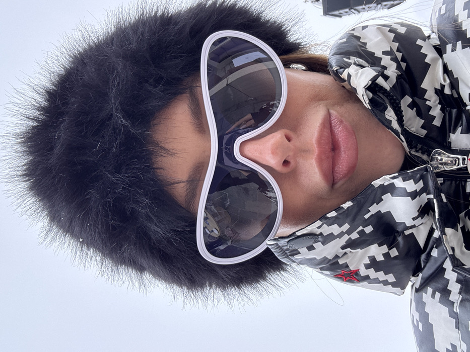 A close-up, low-angle selfie captures an individual in Courchevel, France, under an overcast sky. The subject, appearing to be an adult female, is wearing a large, black, fluffy faux-fur hat or hood, with visible snowflakes on the fur. Oversized, white-framed, dark reflective sunglasses obscure the eyes; reflections in the lenses show parts of a ski resort environment, including structures and possibly other individuals. The subject's nose, mouth with full lips, and chin are visible. They are wearing a black and white geometric, pixelated pattern puffer jacket with a high collar, featuring a red embroidered star on the left side of the collar and a silver-toned zipper with a red detail on the pull tab. The individual is looking directly towards the camera.