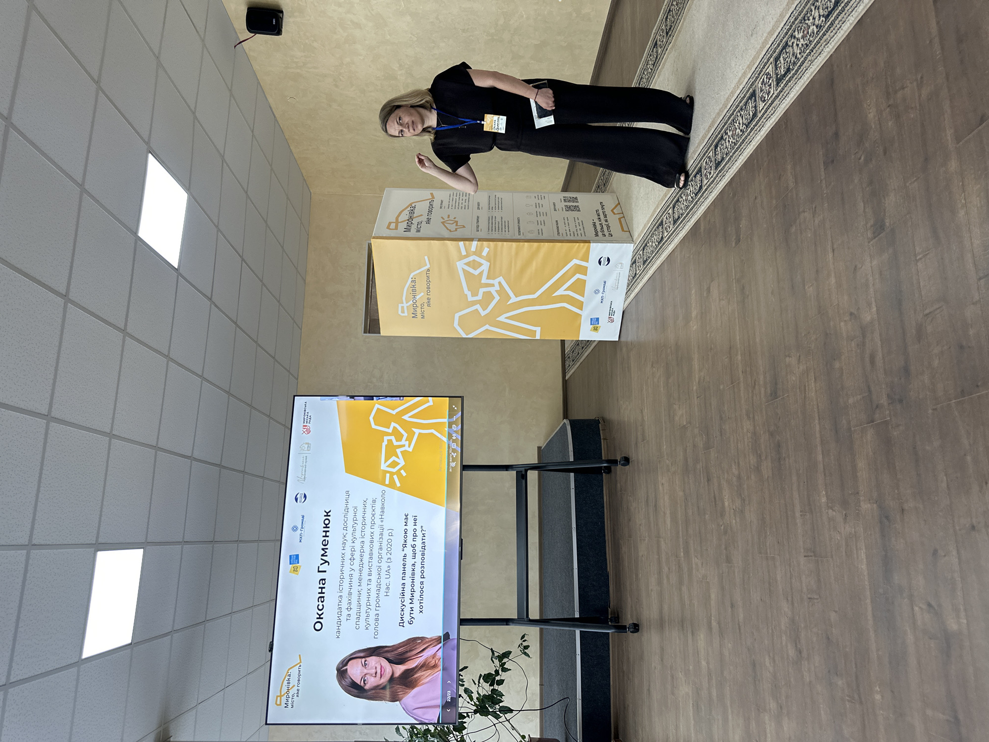 A woman stands to the right of a large, yellow display board, which features Ukrainian text and graphic designs. The woman is wearing a black outfit and a lanyard with an ID card. She is holding a dark object in her left hand. To her left, a large screen displays a presentation about "Oksana Gumeniuk," a candidate of historical sciences, researcher, and manager. The screen also shows the title of a discussion panel: "Myronivka - a city that speaks." In the background, the room has beige walls and a tiled ceiling with fluorescent lighting. The floor is covered with dark wooden planks and a patterned rug. The overall scene suggests an indoor event or presentation related to the city of Myronivka, Ukraine.