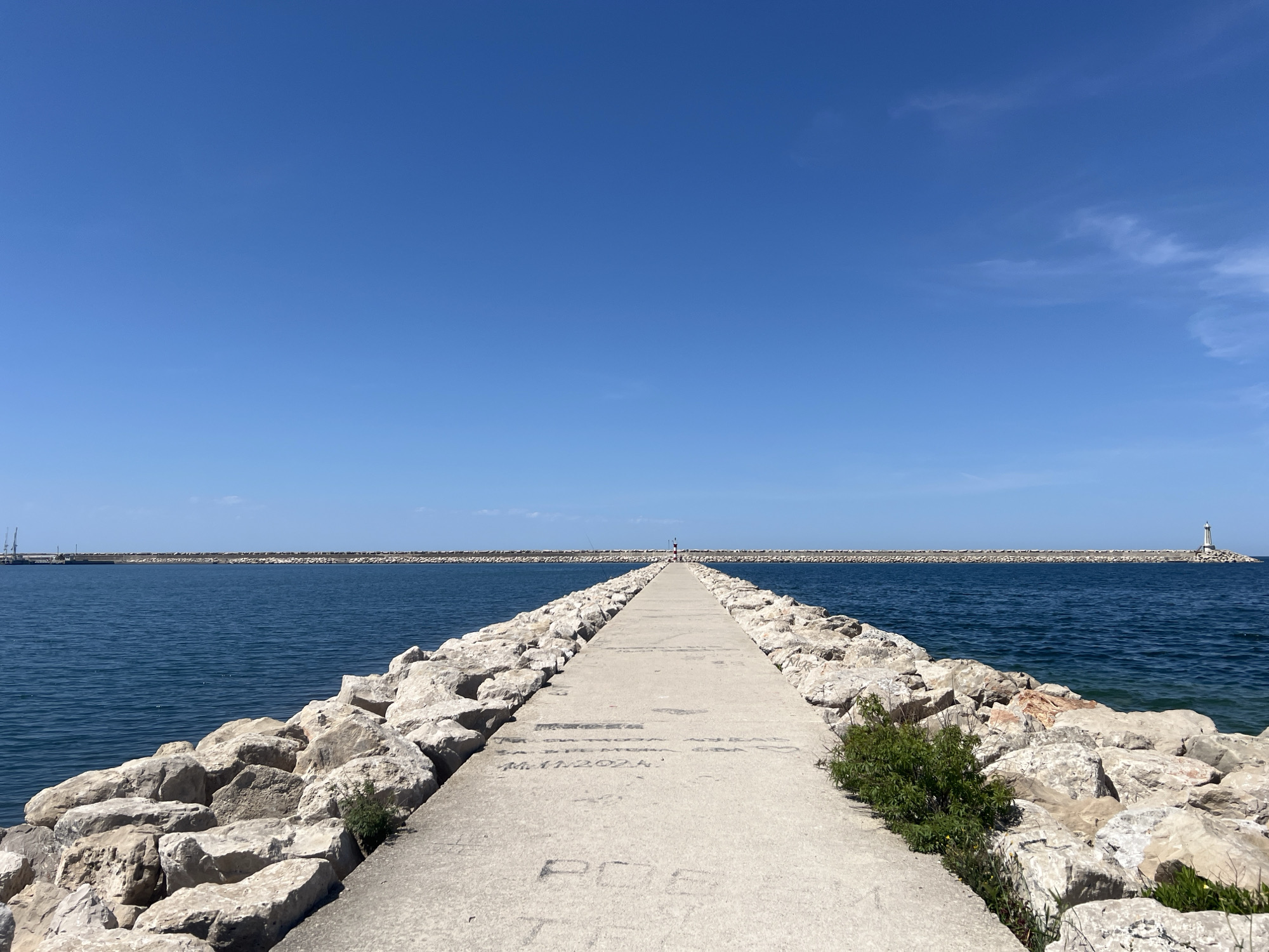A long, straight concrete pier, flanked by large, light-colored rocks, extends into a calm, deep blue sea under a clear, bright blue sky. The pier leads towards the horizon, where a small, distant figure is visible at its end. In the far distance, another rocky breakwater runs horizontally, with a white lighthouse standing prominently at its rightmost end. On the far left horizon, industrial cranes suggest a port area. The water is placid, reflecting the vivid blue of the sky.
Small patches of green vegetation grow between the rocks adjacent to the concrete path, adding a touch of life to the otherwise man-made structure. The weather is sunny and clear, indicating a pleasant day, likely mid-morning or early afternoon, given the bright, direct light. The scene conveys a sense of tranquility and openness.
Several faint inscriptions are visible on the concrete surface of the pier. One prominent, but faded, inscription near the foreground reads "POS". Further down the path, other scribbled texts include what appears to be a date, "M.11.2024", and above it, another date "DA. 17.2024". Other text is too indistinct to read clearly. This location is identified as Bar, Montenegro.