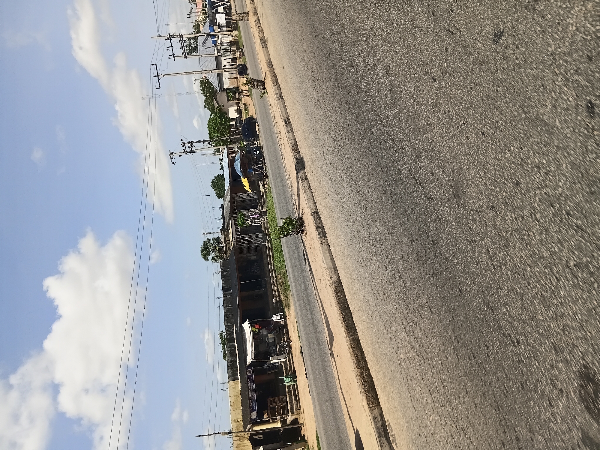 The image depicts a street scene in Jalingo, Nigeria, under a bright sky with scattered white clouds. A paved road occupies the foreground, with a sandy shoulder and a raised curb along its edge. To the left of the road, a row of low-rise buildings with corrugated metal roofs appears to house shops. One shop sign reads "G-AGRO TECH. NIG. LTD." and displays a logo. In front of the shops, there are various objects, including shelves, a bicycle, a wheelbarrow, and what seem to be market stalls with umbrellas. Further down the street, a dark-colored car is parked. Utility poles with electrical wires are visible throughout the scene. The environment appears to be an urban or semi-urban commercial area, possibly a market street.